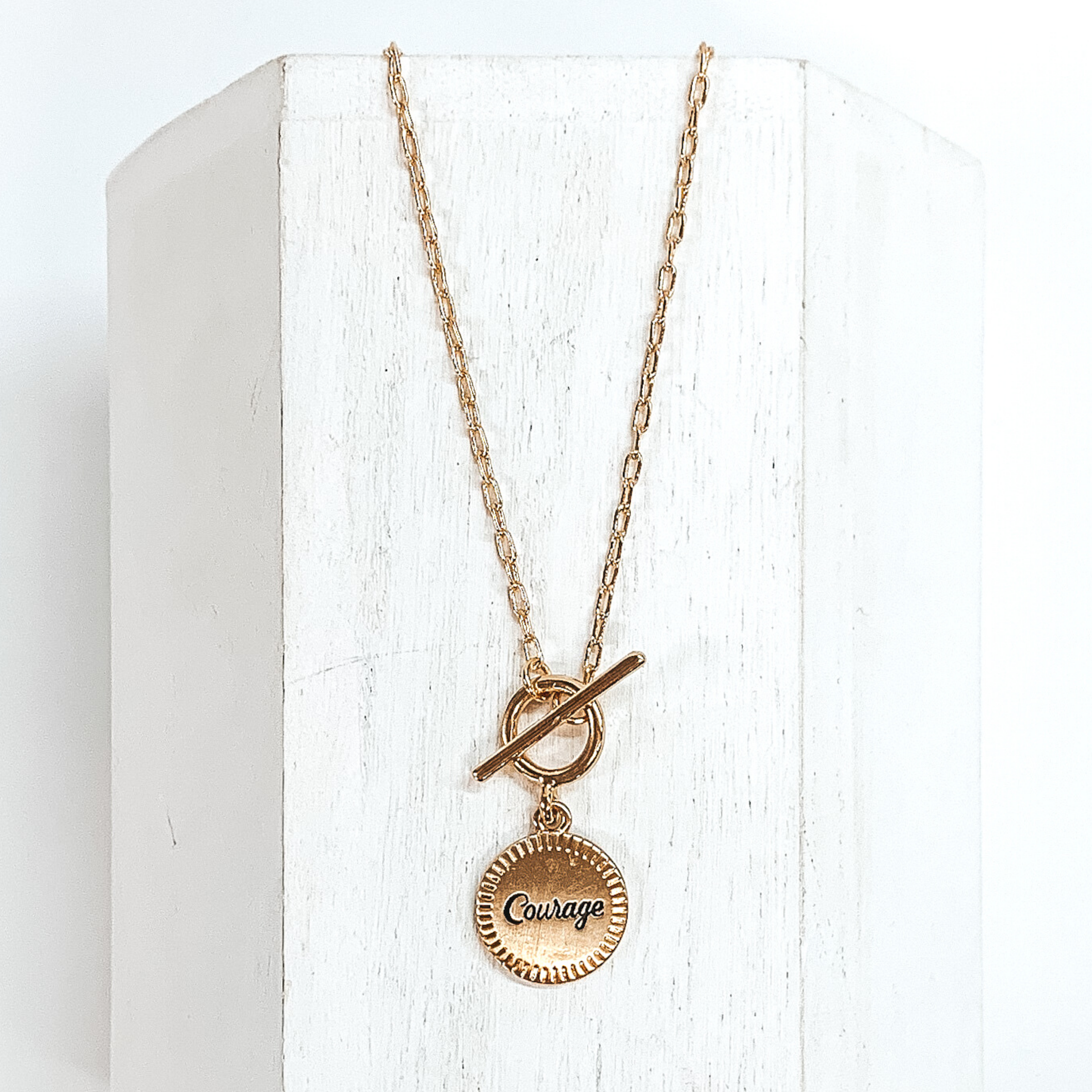 This is a gold chained necklace with a front toggle clasp that has a hanging circle pendant. In the center of the pendant the word "Courage" is engraved in cursive. This necklace is pictured laying on a white block on a white background.