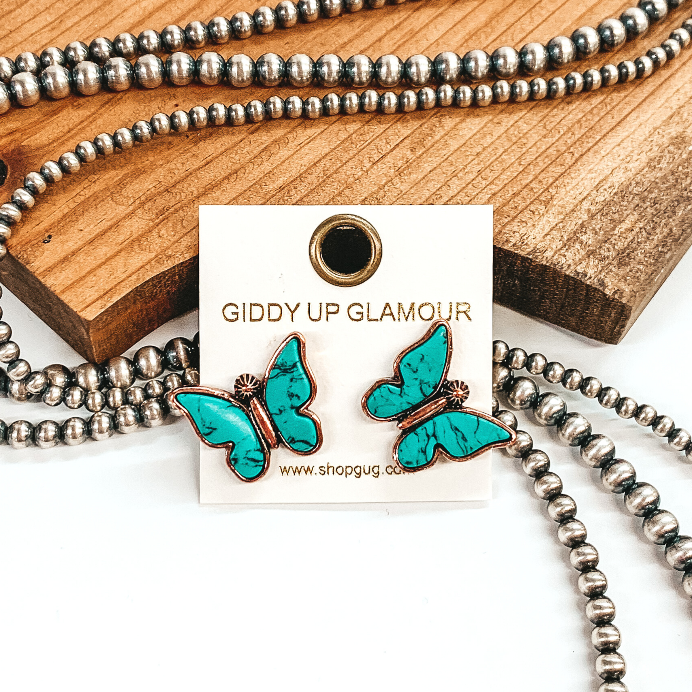 Copper post stud earrings shaped like a butterfly with turquoise stones for wings. These earrings are pictured in front of brown wood with some silver pearls laying behind the earrings.