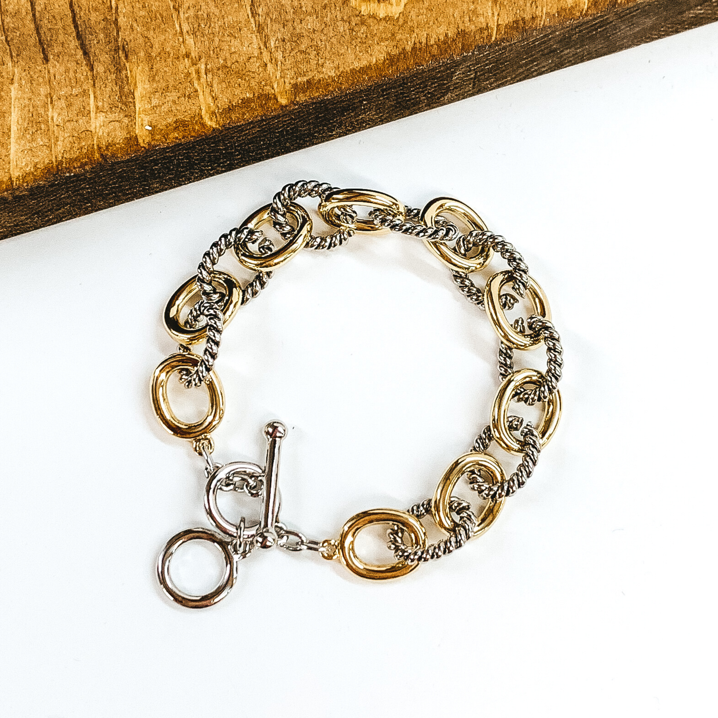 Small silver, twisted oval chain linked with smooth, gold chain links bracelet. This bracelet has a toggle clasp. This bracelet is pictured on a white background.