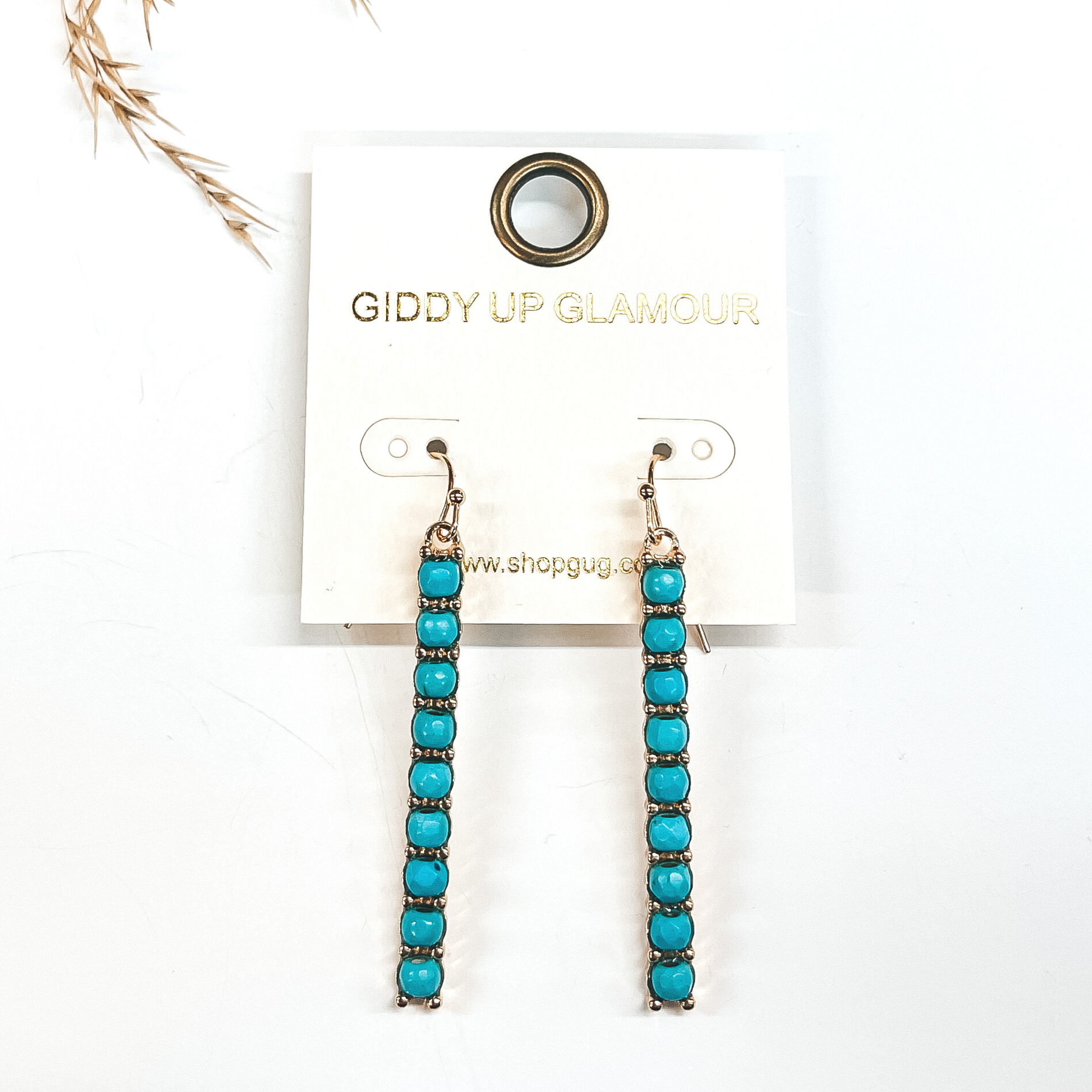 Gold rectangle drop earrings with turquoise colored beads all the way down.  Taken on a white background with a brown plant in  the back as decor.