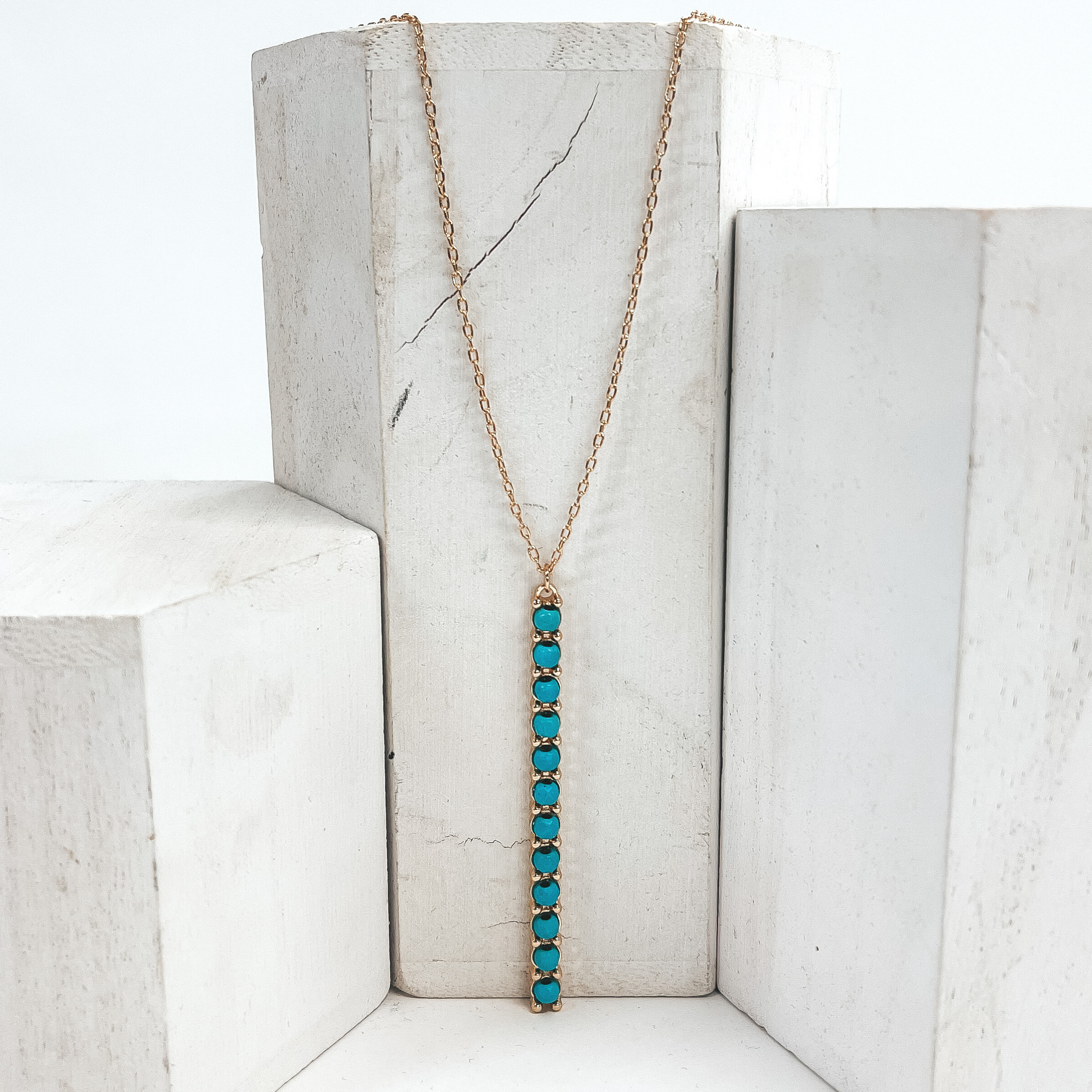 This is a 30 inch adjustable gold necklace with  a gold 2.75 inch bar pendant with turquoise   beads. This necklace is pictured laying on a white  block on a white background with one white block on  each side.