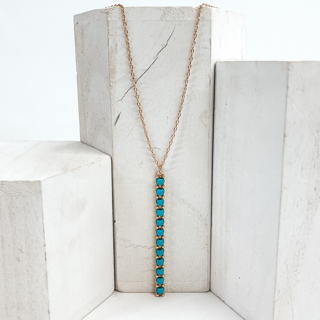 This is a 30 inch adjustable gold necklace with  a gold 2.75 inch bar pendant with turquoise   beads. This necklace is pictured laying on a white  block on a white background with one white block on  each side.