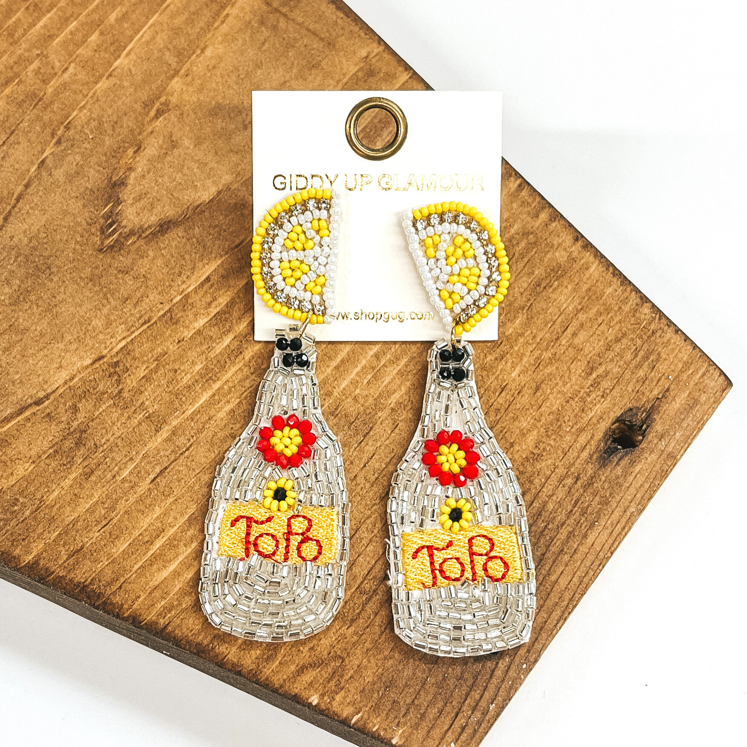 Beaded lemon stud earrings with a hanging bottle pendant. The bottle pendant is covered in clear crystals with a red and yellow beaded flower. It also includes a yellow stitched area that has the word "ToPo" stitched in red. These earrings are pictured on a dark brown object on a white background.