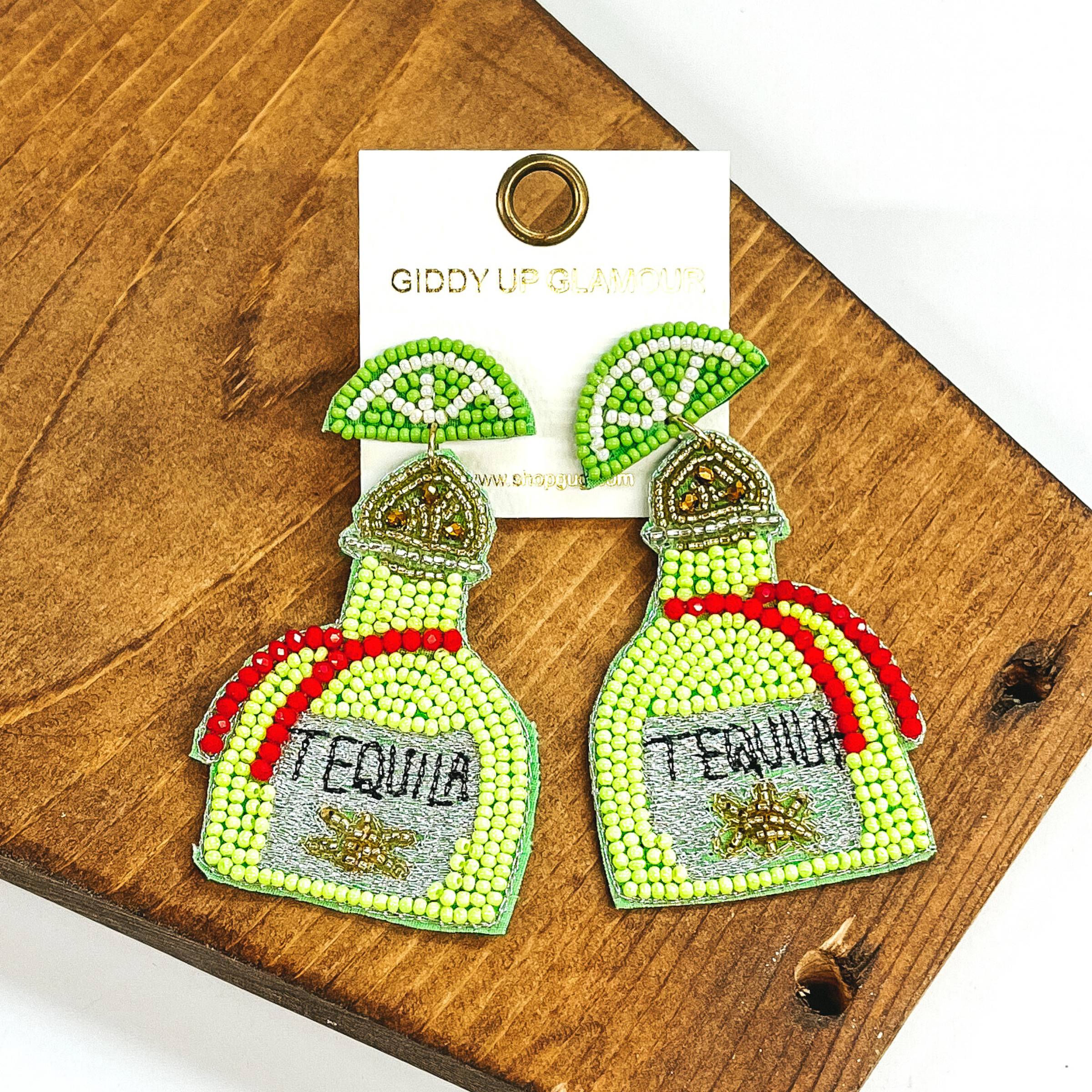 Light green beaded lime stud earrings with hanging beaded bottle. The bottle has a gold beaded top with a lime green beaded body with red beadedstripes. The center on the bottom is grey stitched with the word "TEQUILA" stitched in black. These earrings are pictured on a dark brown object on a white background.