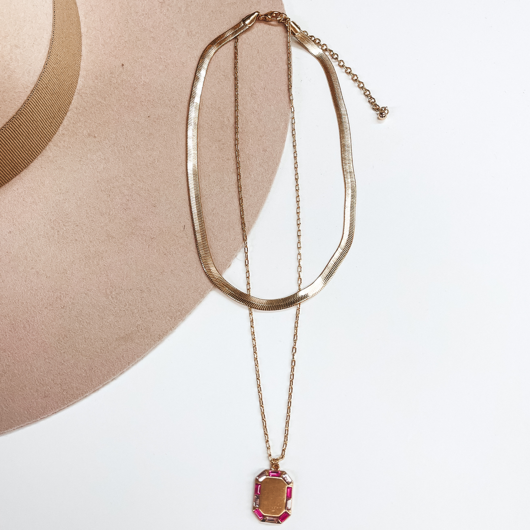 Gold double strand necklace, the small strand is a  snake chain and the longer strand is a regular  small chain. The longer strand has a rectangular gold  pendant with crystals around in light pink and hot pink. This necklace is pictured on a white and  beige background.