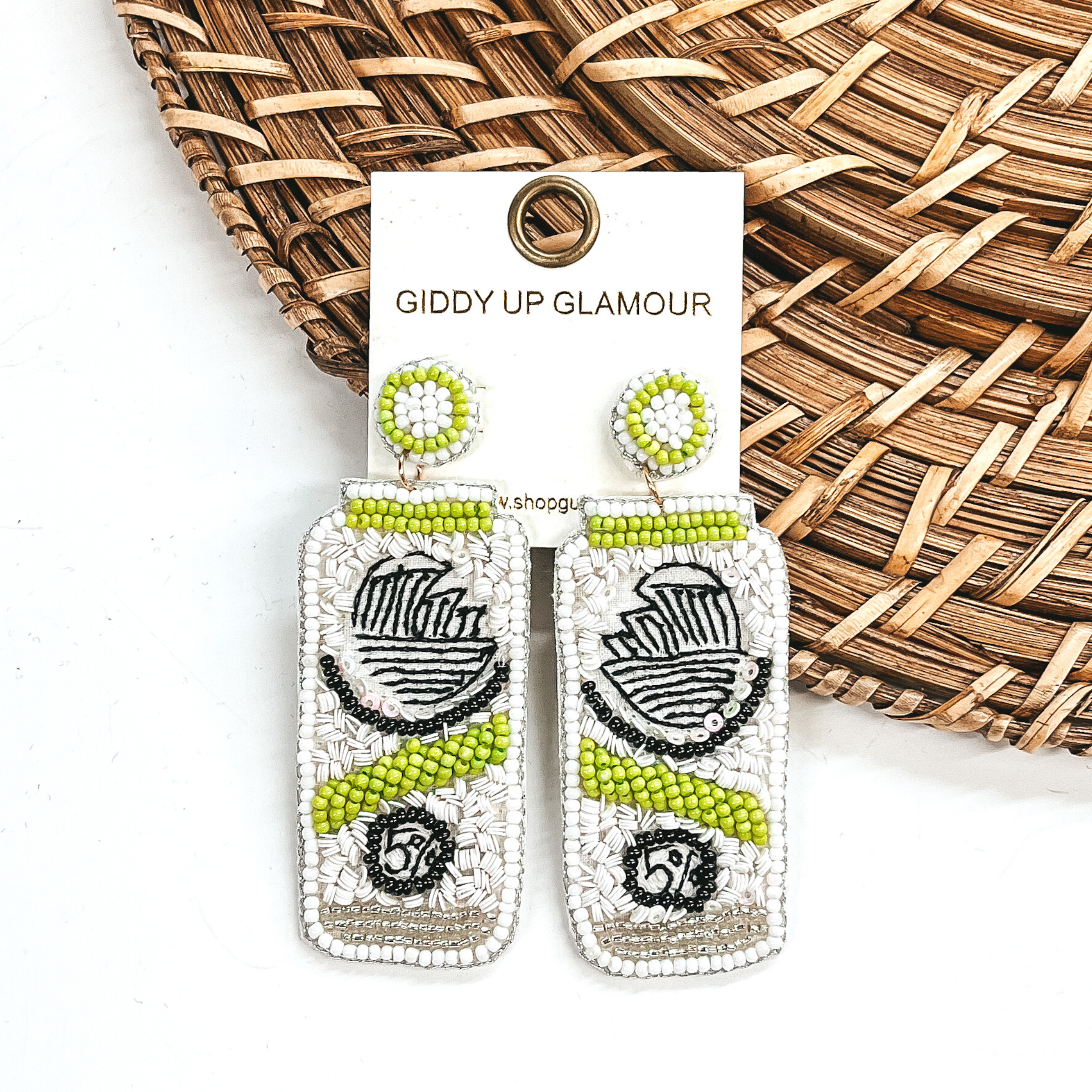 White beaded can earrings with lime green accents that look like White Claw cans. These earrings are pictured laying partially on a basket weave material on a white background. 