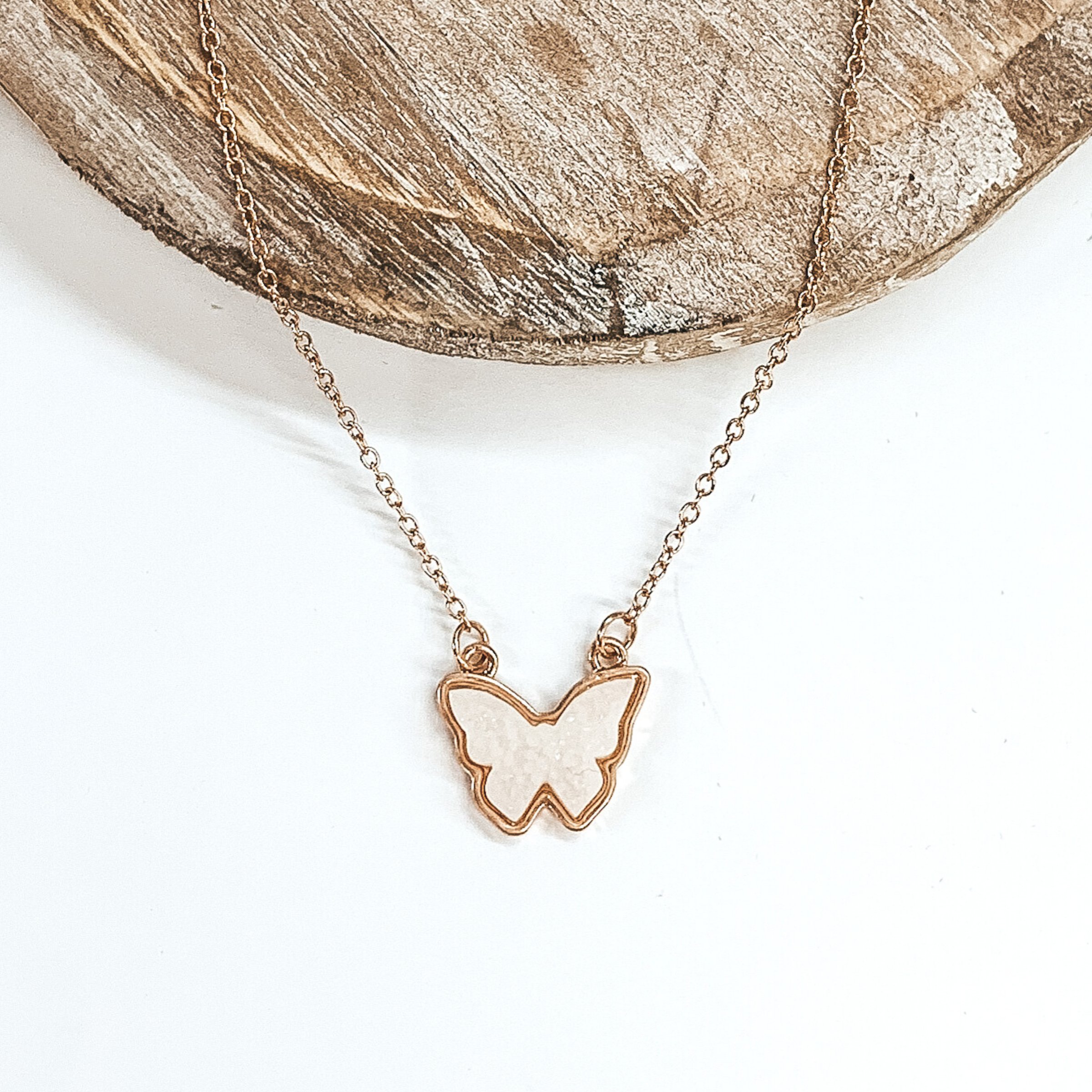 Tiny gold chained necklace includes a white, druzy butterfly pendant that is outlined in gold. This necklace is pictured on a white background with a tan piece of wood at the top of the picture. 