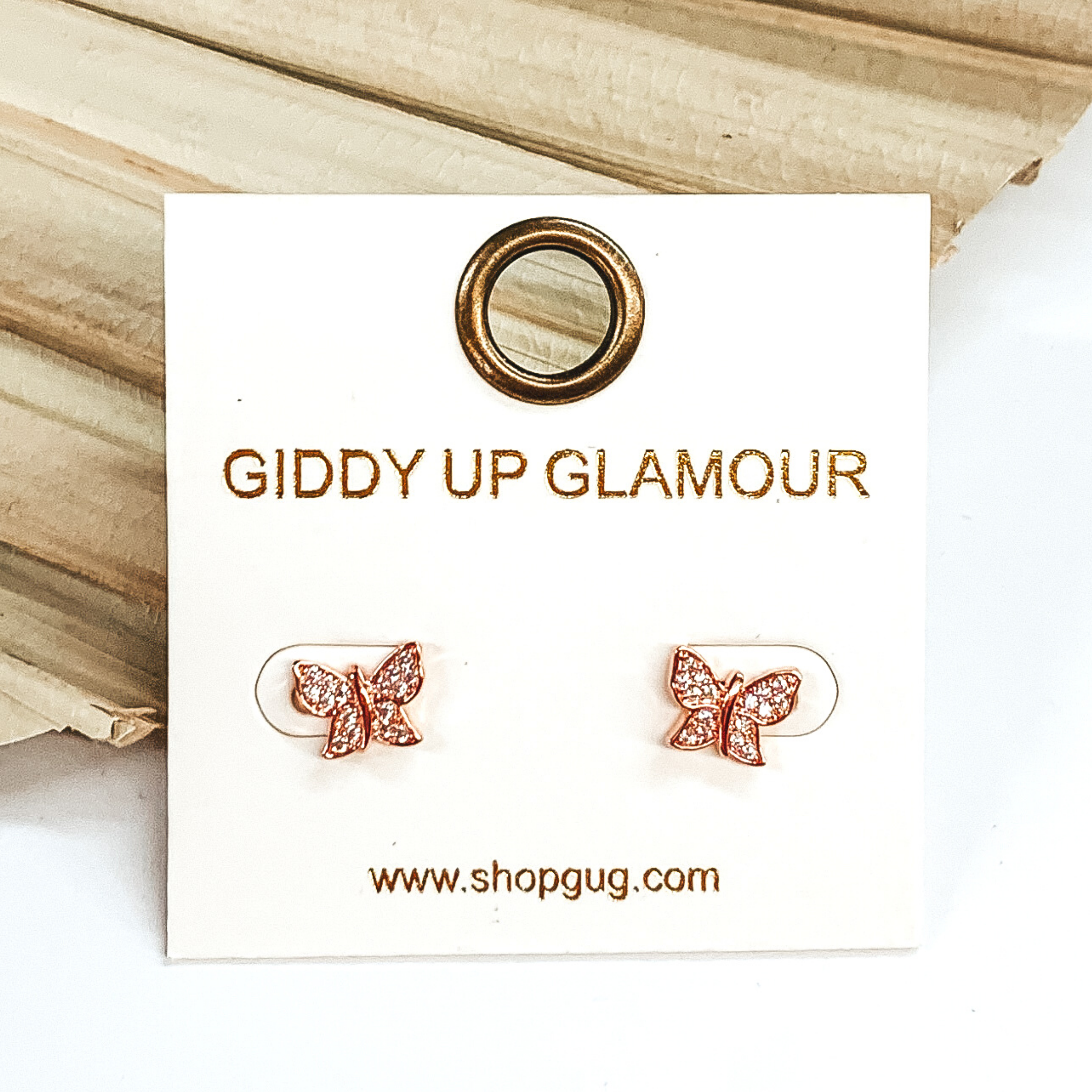 Small, rose gold butterfly stud earrings with clear crystals. These earrings are pictured laying on a green palm leaf on a white background.