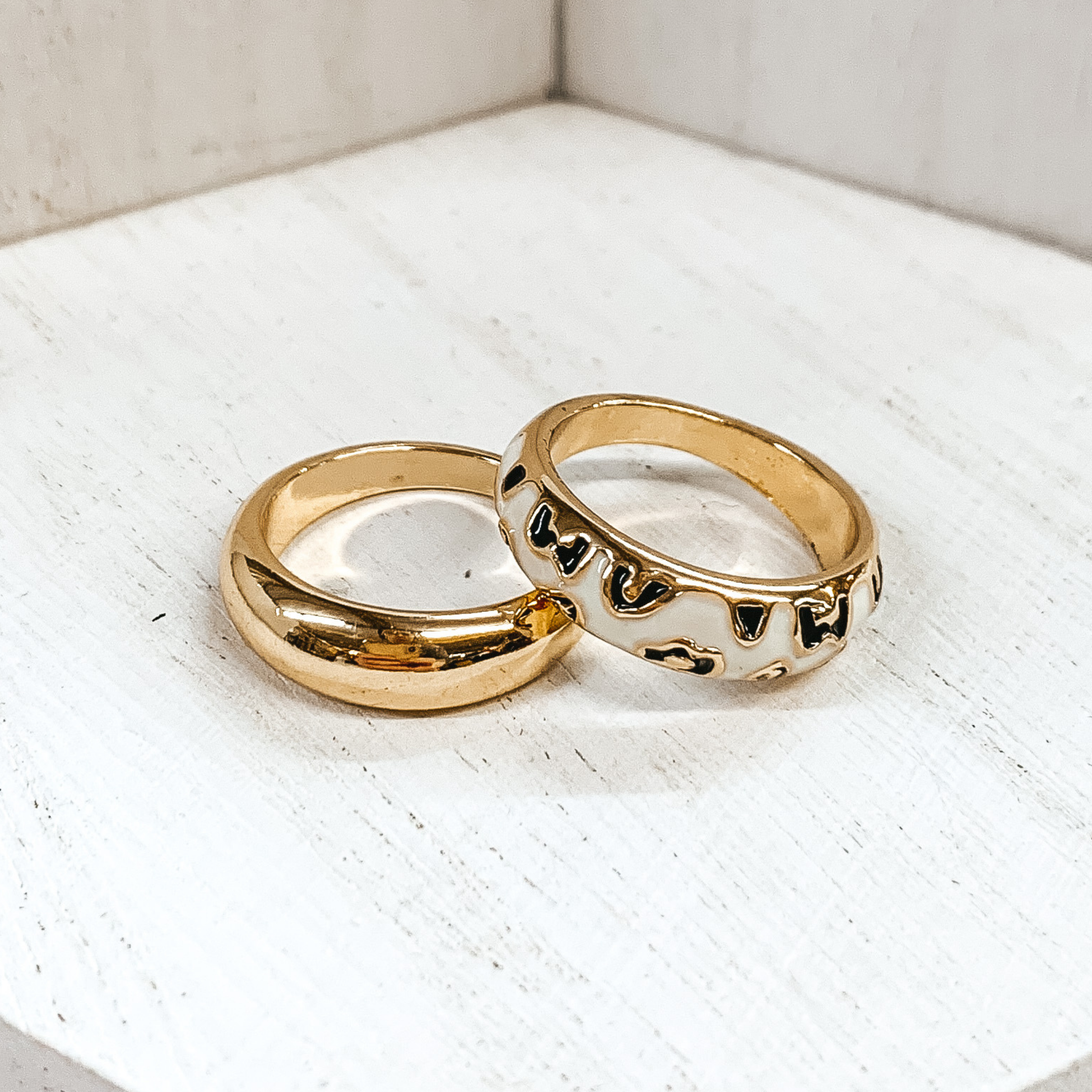 Set of two thick, gold rings. One ring is plain, while the secong ring has a white colored part with black leopard print. These rings are pictured on a white background. 