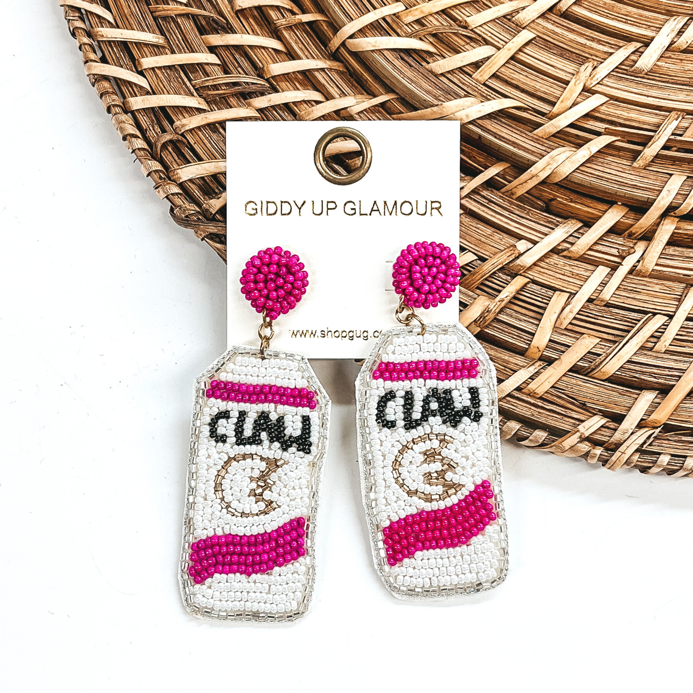 White beaded can earrings with pink accents that look like White Claw cans. These earrings are pictured laying partially on a basket weave material on a white background.
