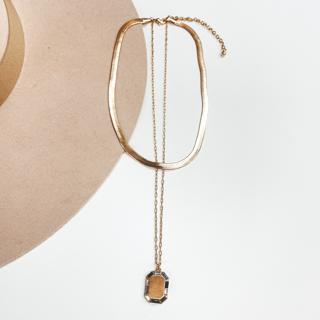 Gold double strand necklace, the small strand is a  snake chain and the longer strand is a regular  small chain. The longer strand has a rectangular gold  pendant with crystals around in black, gray, and  clear. This necklace is pictured on a white and  beige background.
