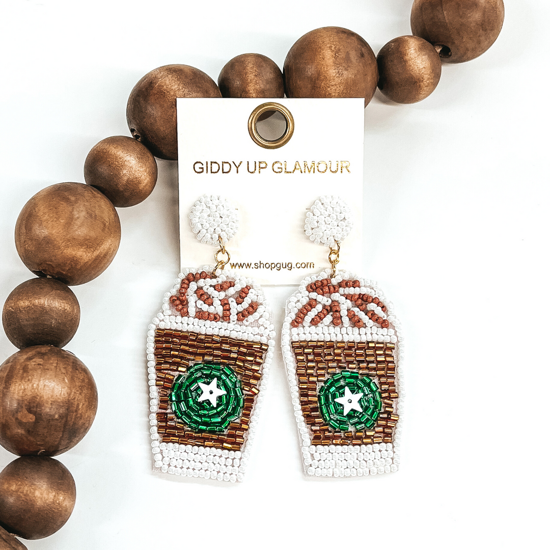 White and bronze beaded cup with green circle with white star. There is also a white and brown beaded whip on the top of the cup. This coffee pendant is hanging from white beaded circle studs. These earrings are pictured in front of dark brown beads on a white background. 