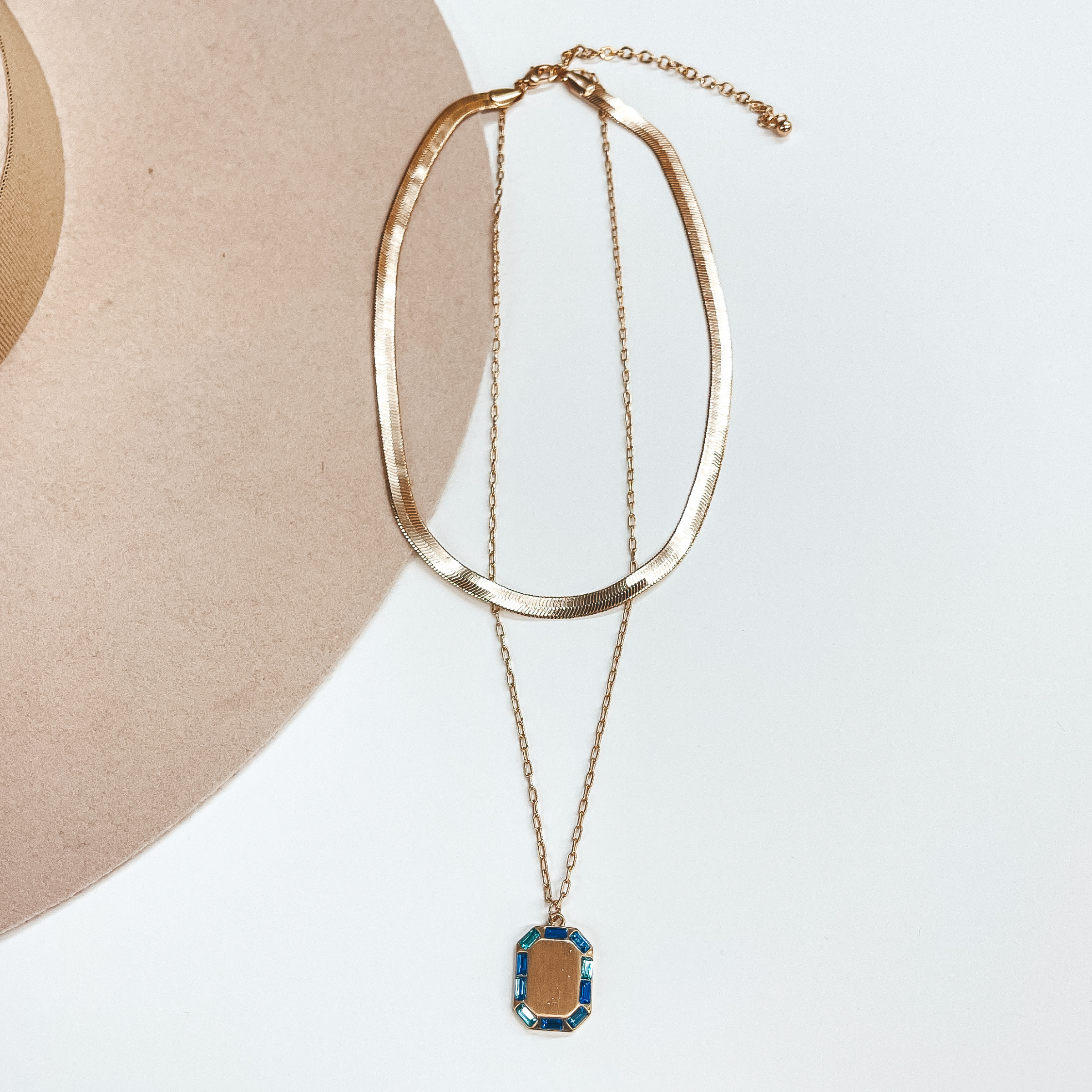 Gold double strand necklace, the small strand is a  snake chain and the longer strand is a regular  small chain. The longer strand has a rectangular gold  pendant with crystals around in blue, light blue,  and teal. This necklace is pictured on a white and  beige background.