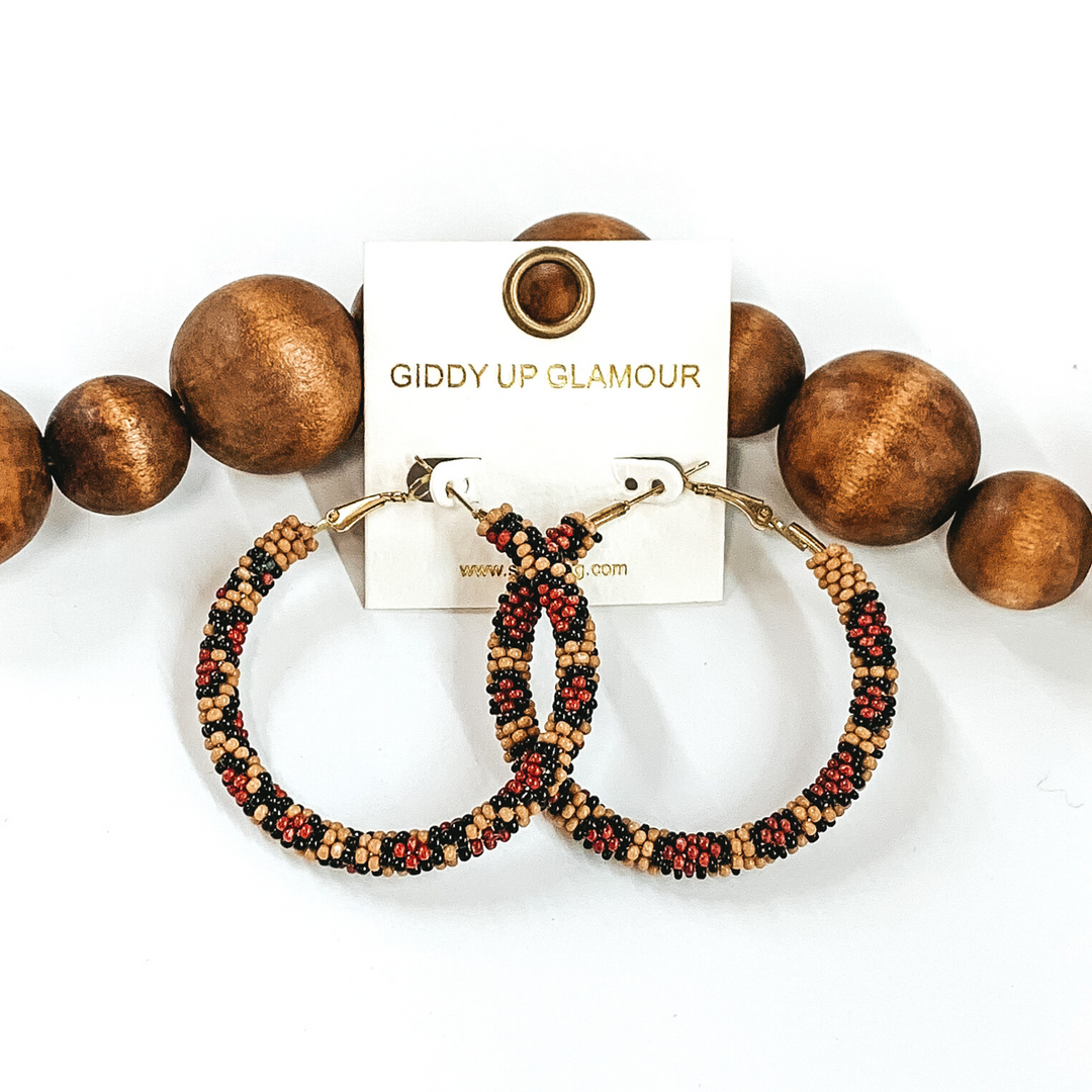 Beaded hoop earrings in leopard print. These earrings are pictured laying in front of dark brown beads on a white background. 