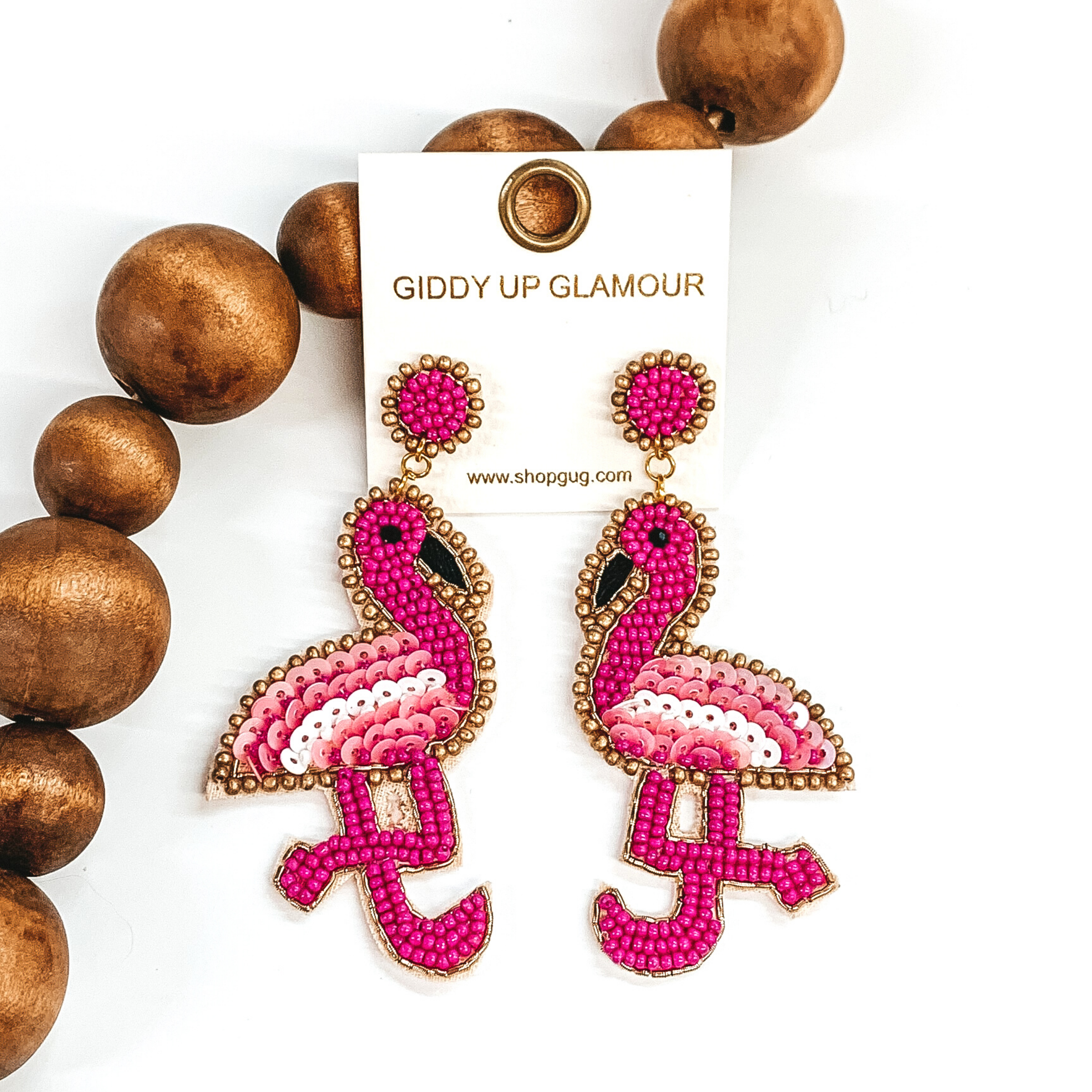 These earrings include circle studs with fuchsia beads and a gold outline. Hanging from the bottom of these studs is a flamingo shaped beaded pendant. These earrings are pictured leaning agasint dark brown beads on a white background. 