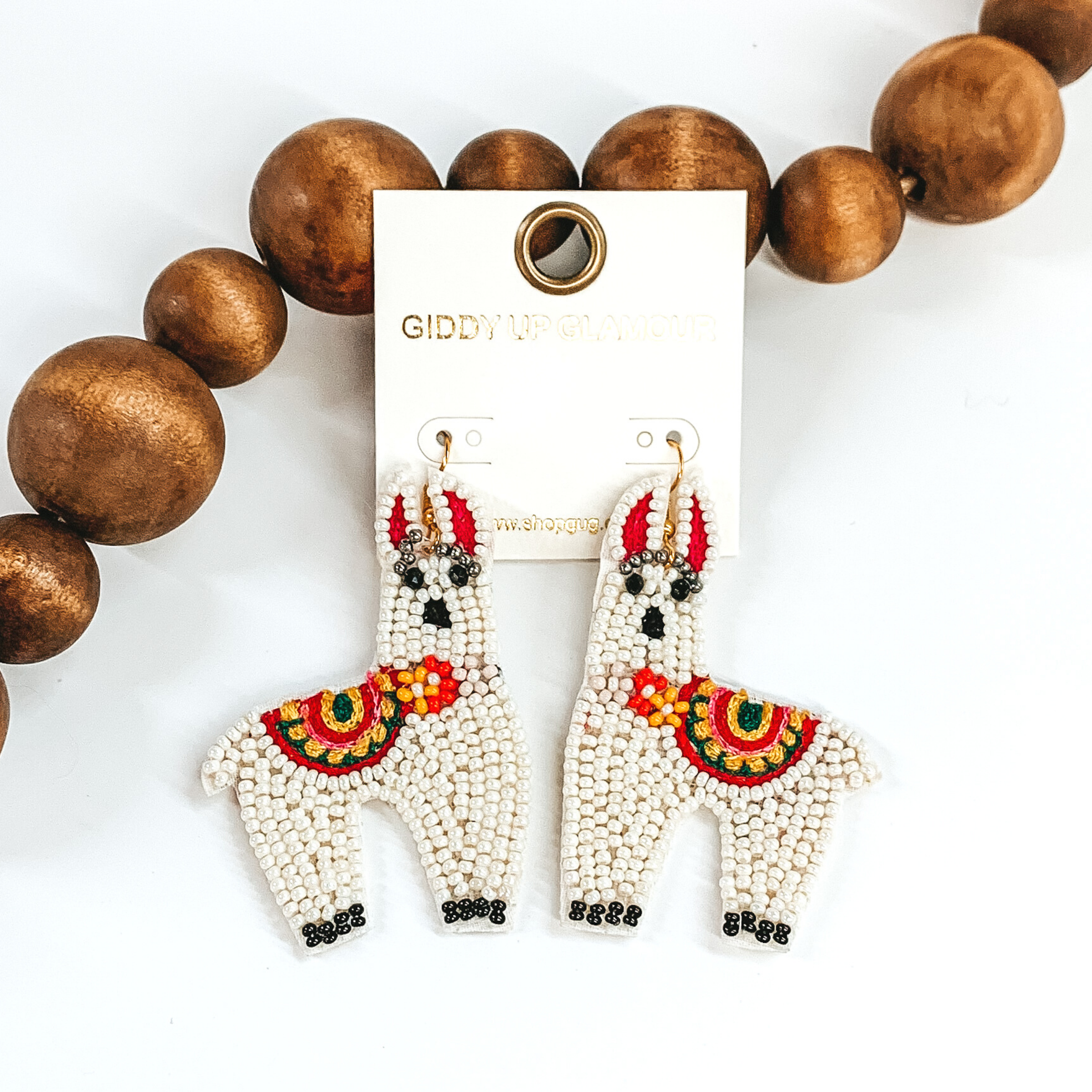 Llama shaped beaded dangle earrings in ivory. These earrings also includes some colorful stitching details. These earrings are pictured on a white background with dark brown beads towards the top of the picture.