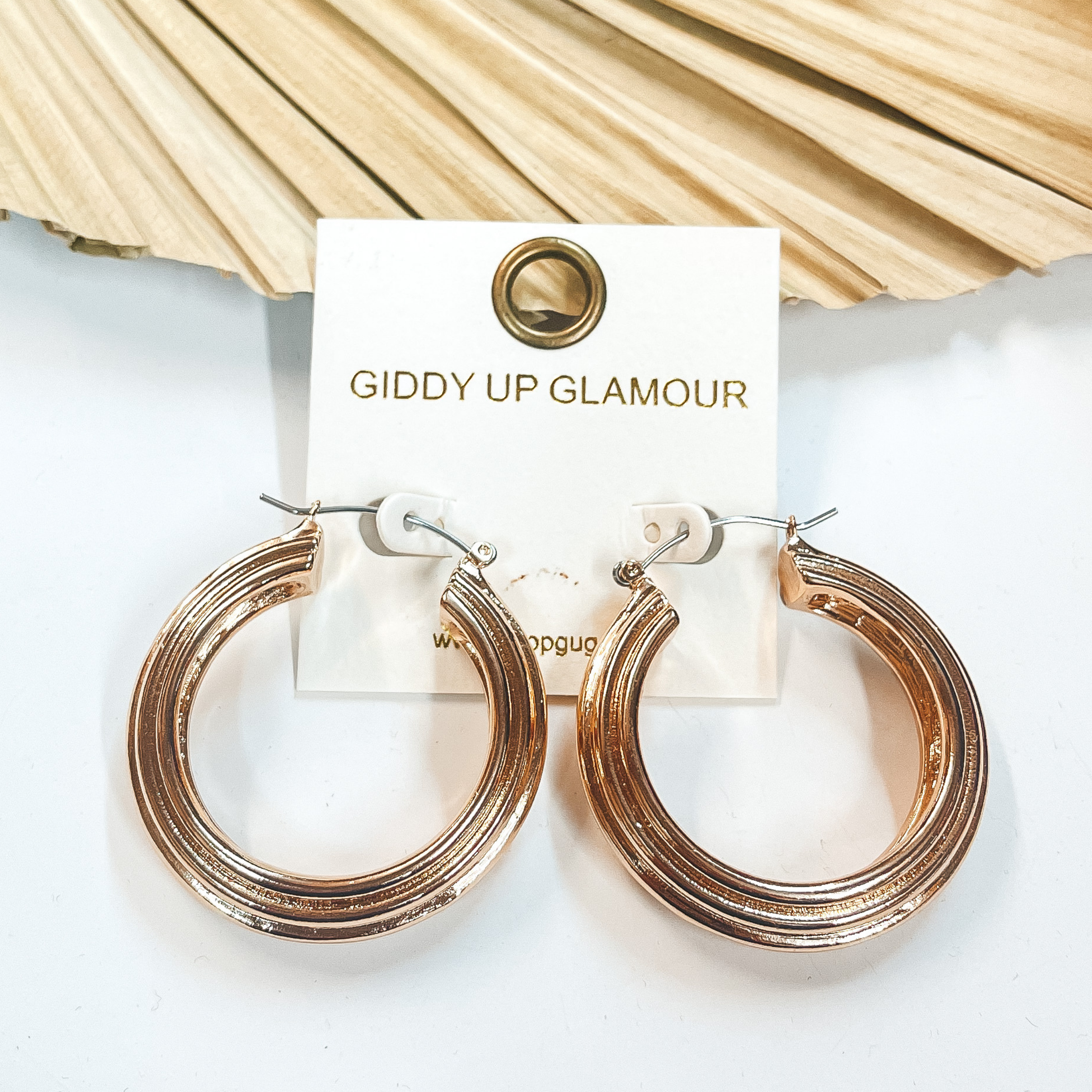 Thick gold hoop earrings with lined texture. Taken  on a white background and leaned up against a  dried up palm leaf.
