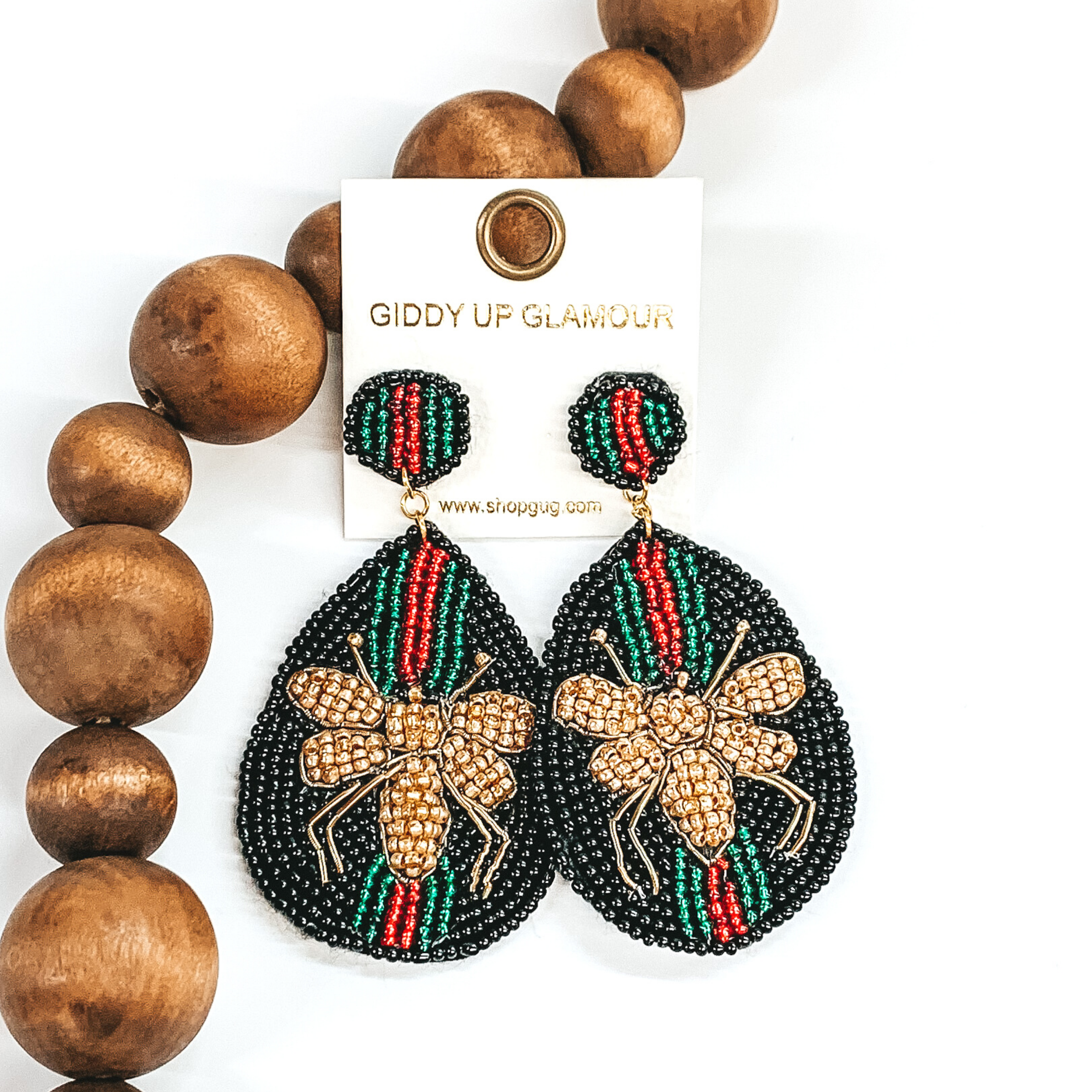Circle beaded studs with a beaded teardrop dangle. The majority of the beads were black with green and red stripes. The center of the teardrop was a gold beaded bee. These earrings are pictured on a white background with dark brown beads. 
