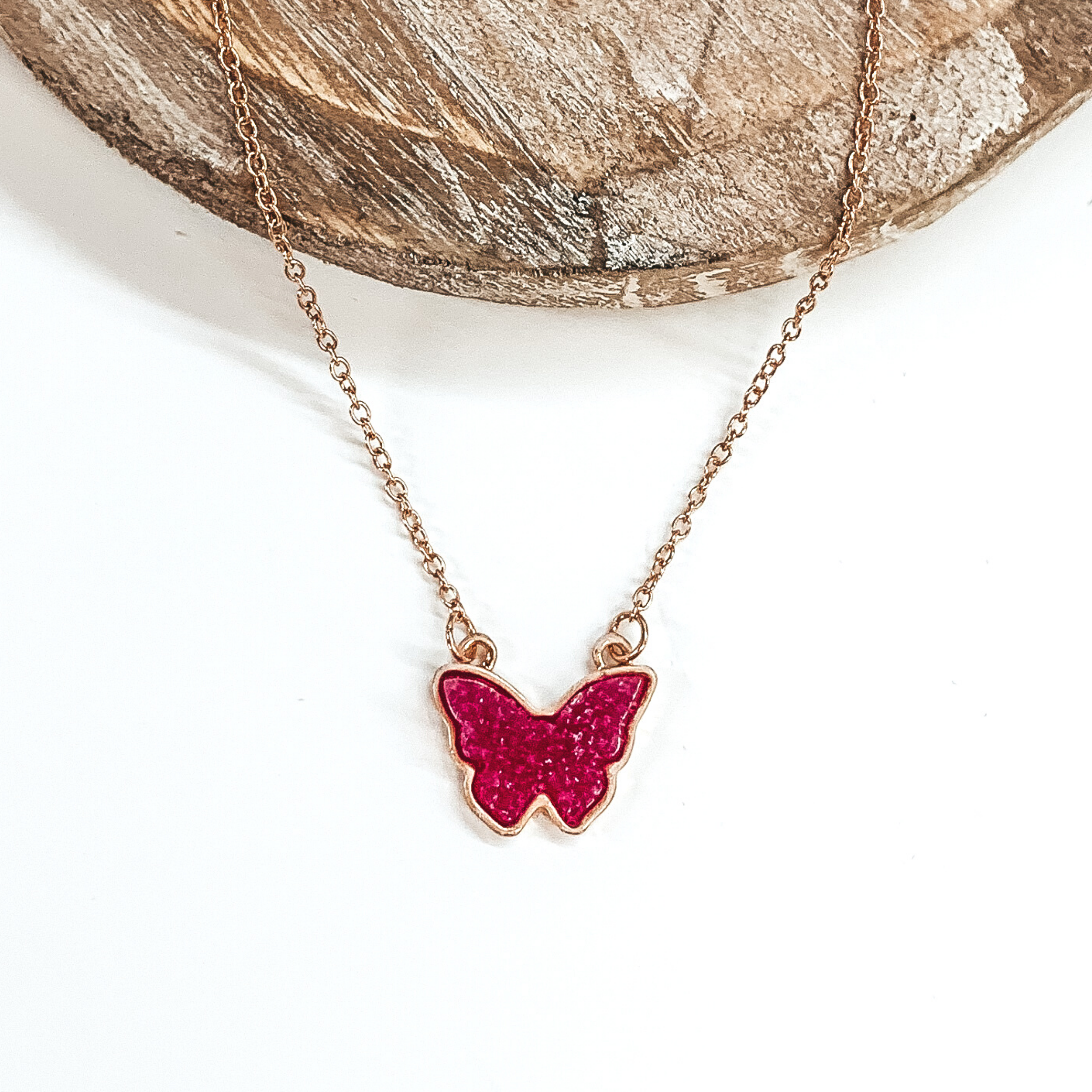 Tiny gold chained necklace includes a fuchsia, druzy butterfly pendant that is outlined in gold. This necklace is pictured on a white background with a tan piece of wood at the top of the picture.
