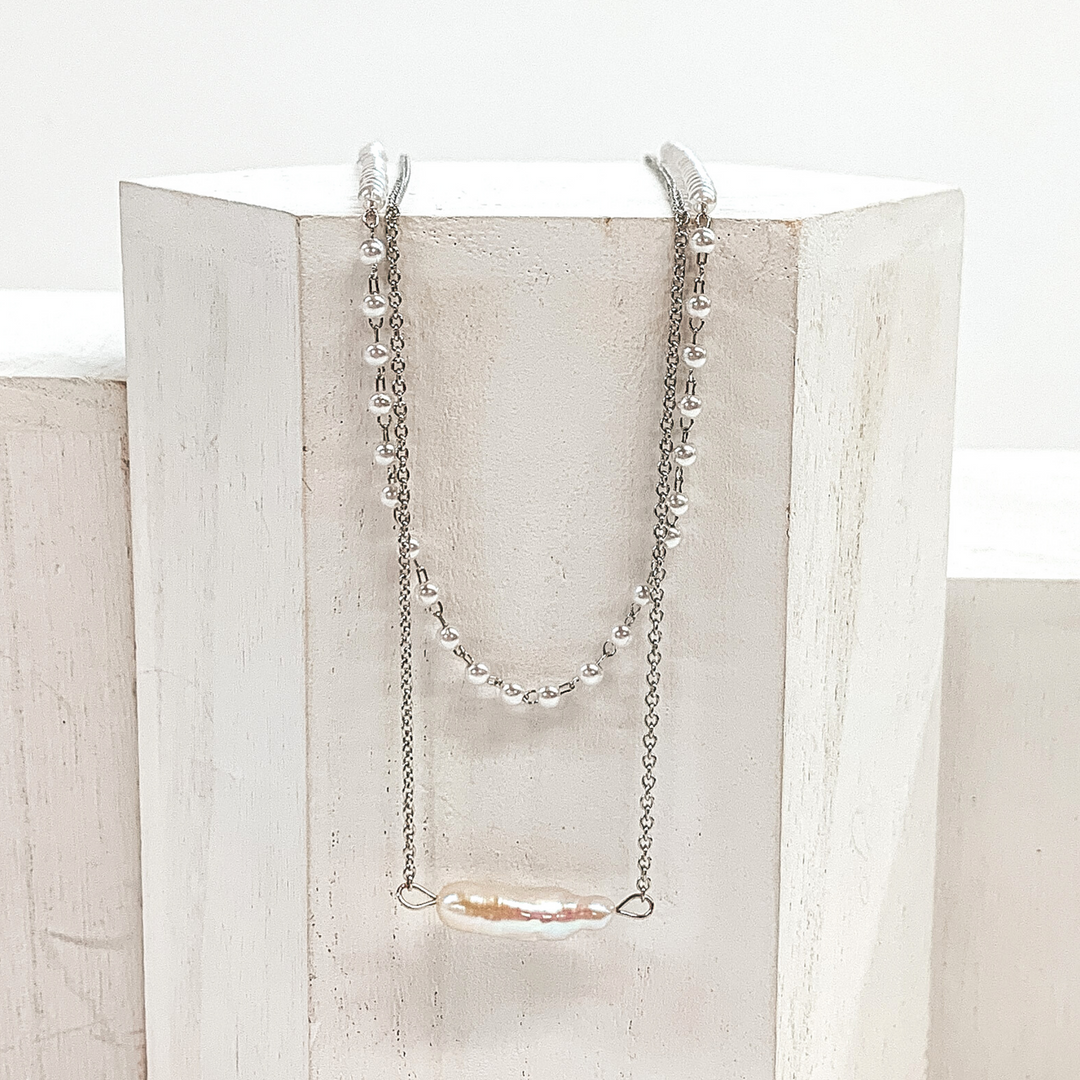 This is a silver, two stranded necklace that includes one silver chained necklace with an irregularly shaped pearl pedant and a pearl beaded necklace. This necklace is pictured laying on a white block on a white background.