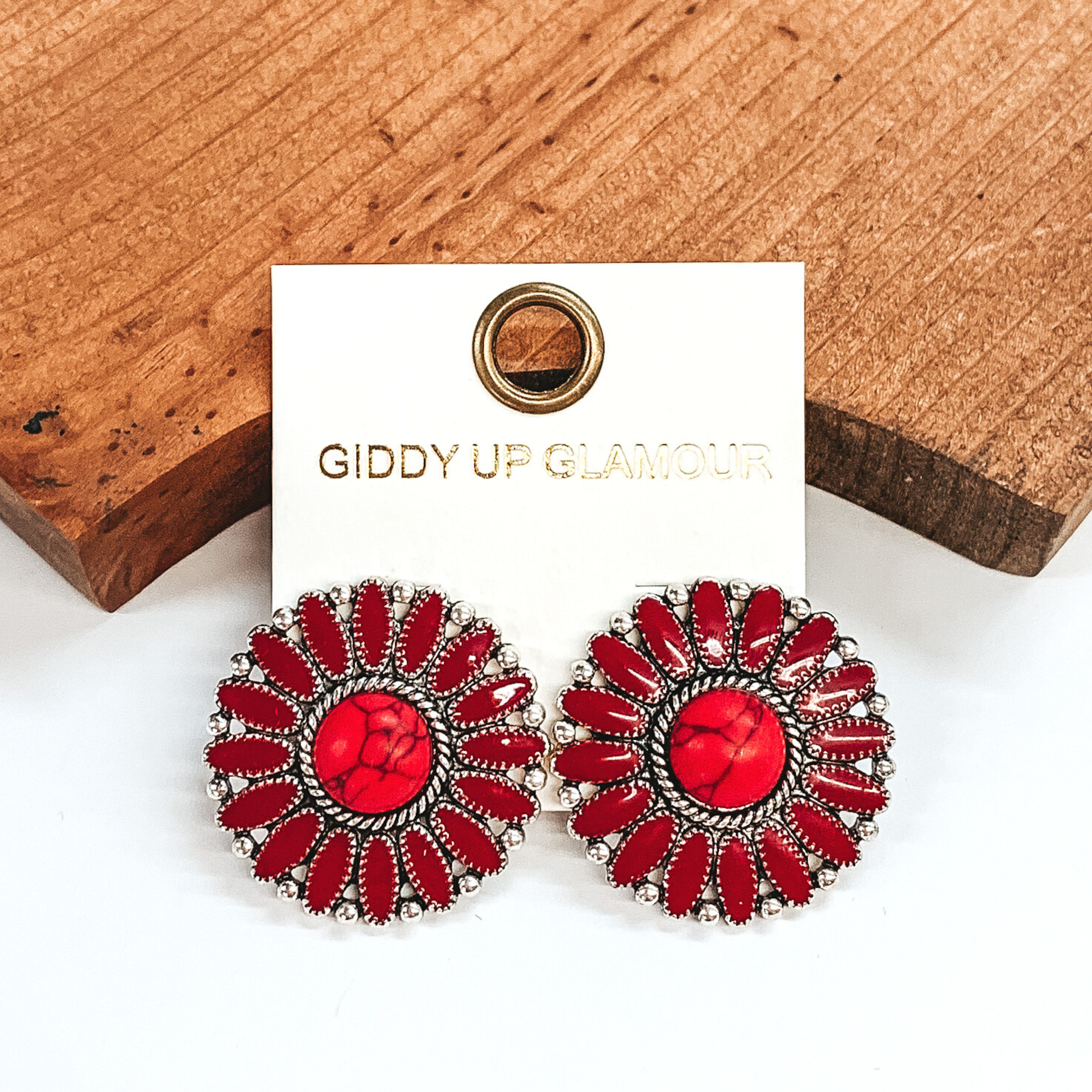 Maroon concho earrings with a center red stone and silver outline. These earrings are pictured on a white earrings holder laying against a dark brown block on a white background.