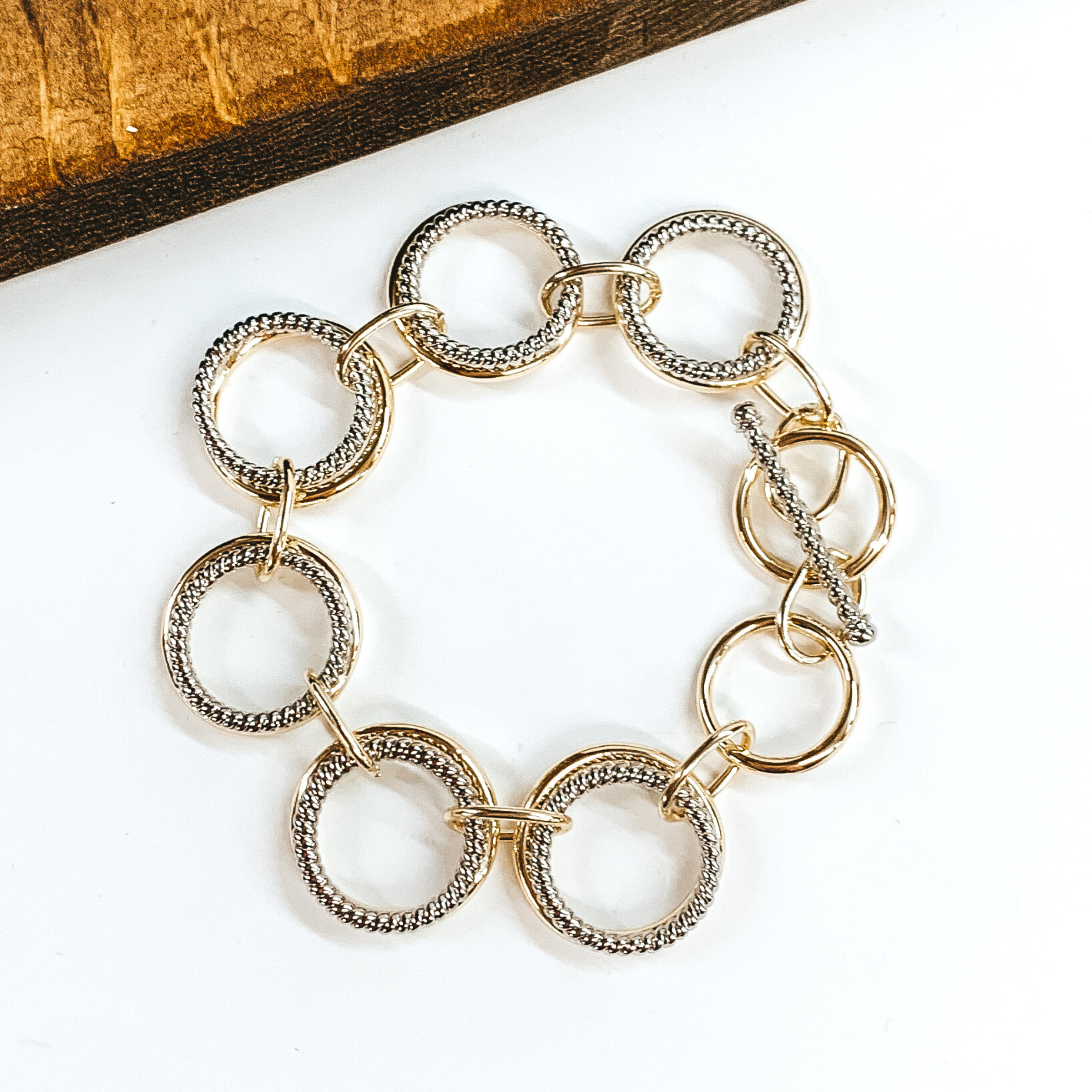 This bracelet has double circle chain link that include a smooth gold circle and a twisted silver circle. These links are connected by thin, gold rectangle chains. This bracelet has a toggle clasp. This bracelet is pictured on a white background.