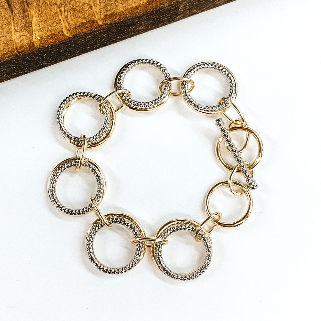This bracelet has double circle chain link that include a smooth gold circle and a twisted silver circle. These links are connected by thin, gold rectangle chains. This bracelet has a toggle clasp. This bracelet is pictured on a white background.