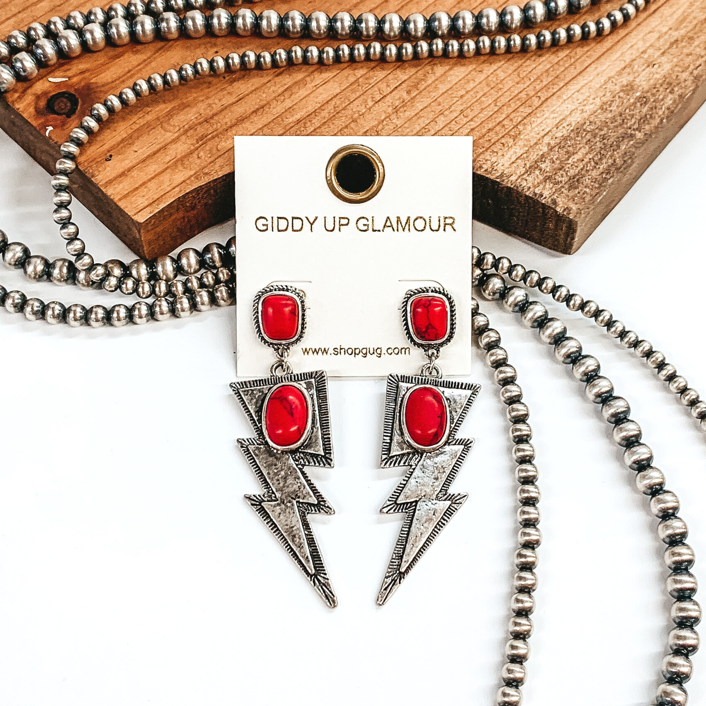 Silver irregular shaped stud earrings with a red colored stone. Hanging from the bottom there is a silver lightning bolt pendant with an oval red stone. These earrings are pictured in front of brown wood with some silver pearls laying behind the earrings. 