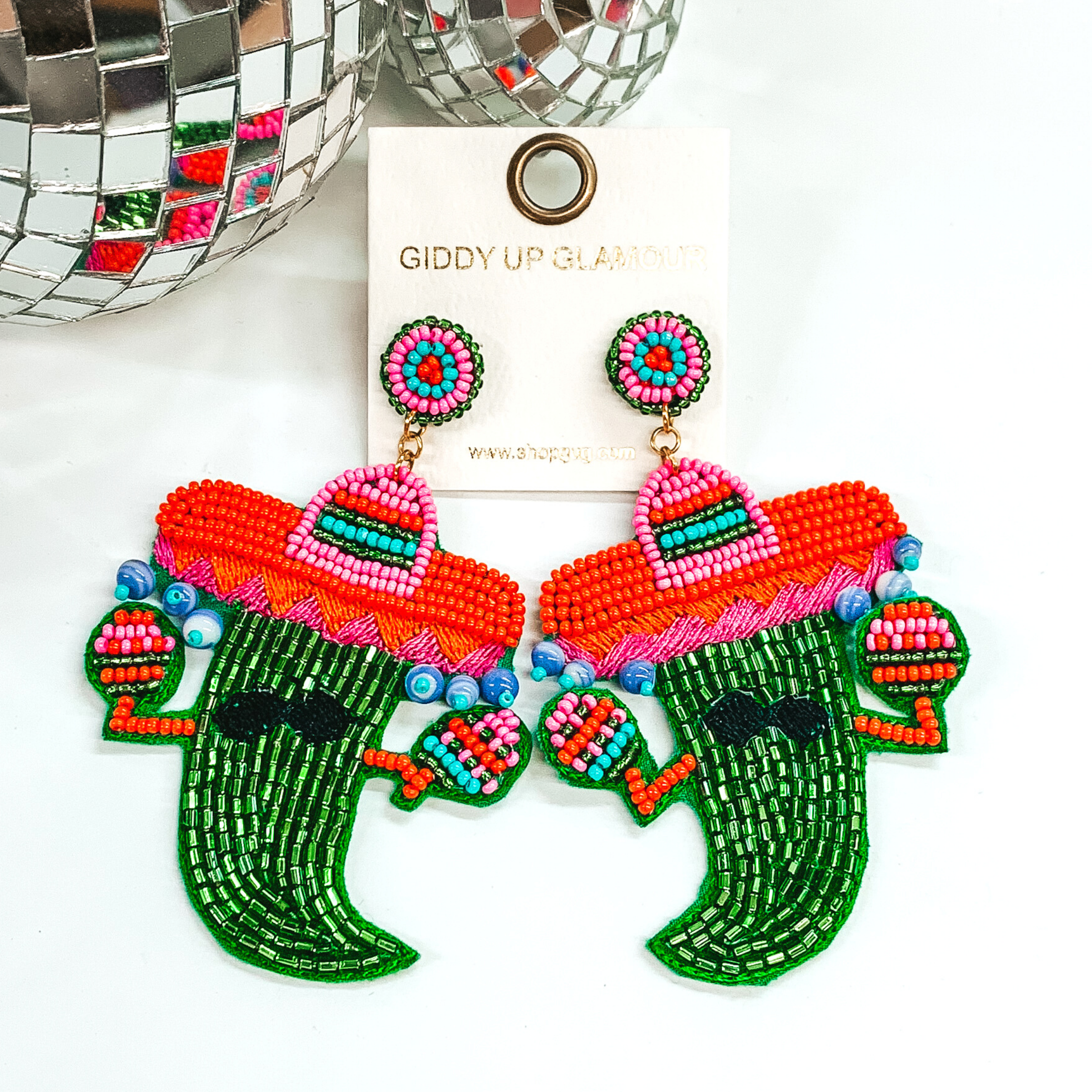 Green chili pepper beaded earrings. This pepper has arms holding colorful maracas, has a black stitched mustache, and a colorful sombrero with beaded tassels. These earrings are pictured on a white background with disco balls in the top left corner. 