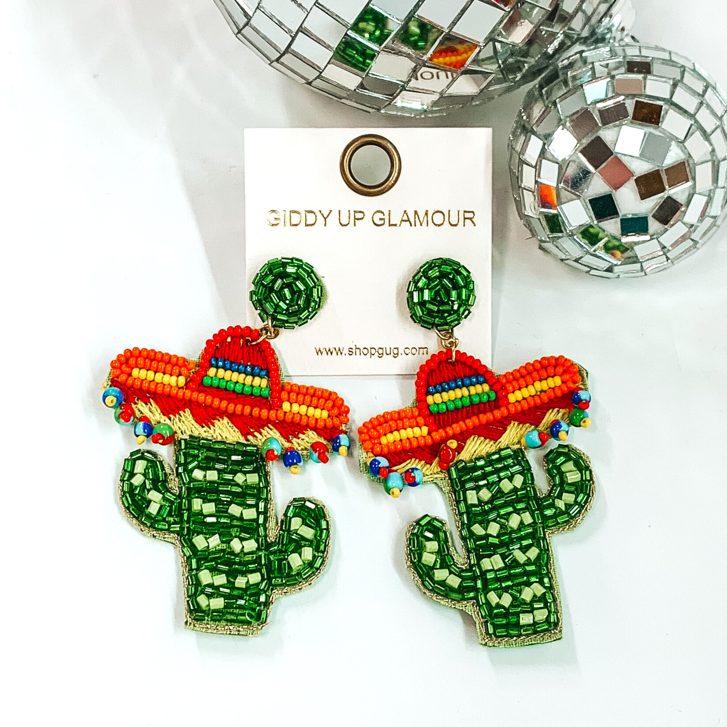 These earrings are beaded cactus earrings in light and dark green. the cactus has a colorful sombrero on the top of the cactus. The cactus pendant is hanging from a green, beaded circle stud. These earrings are pictured on a white background with disco balls in the top right corner.