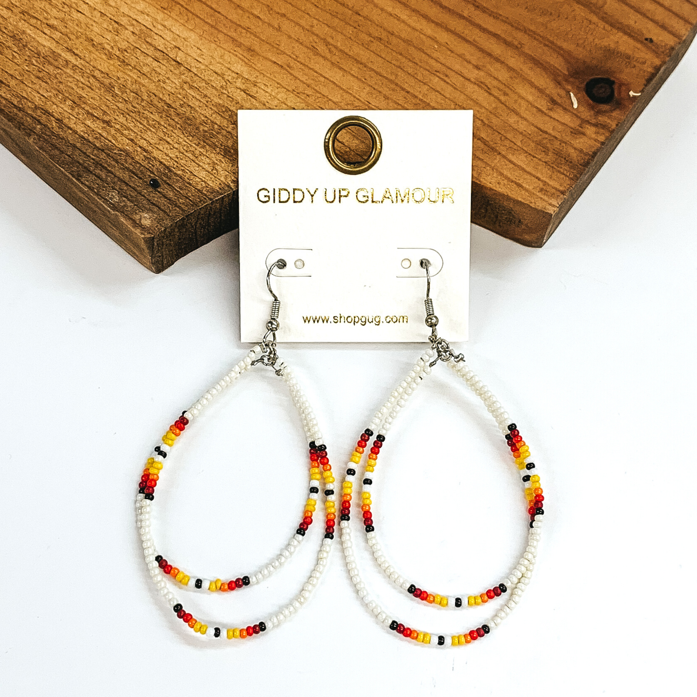 Double layered, hanging beaded teardrop earrings. The majority of the beads are white with three segements including maroon, red, yellow, white, and black beads. hese earrings are pictured on a white background in front of a dark piece of wood. 