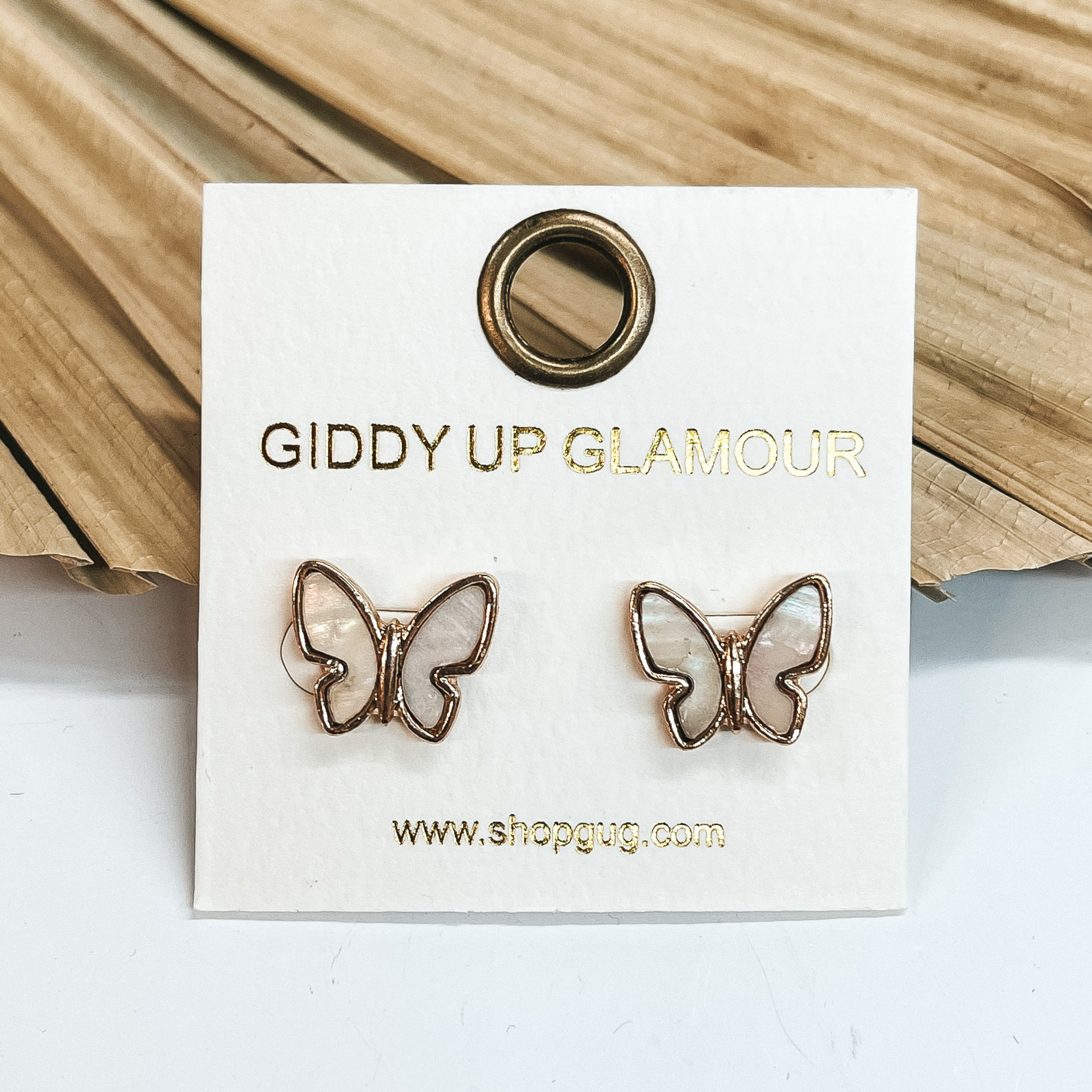 These are mother of pearl butterfly earrings with  a gold setting. Taken on a white background and leaned up against a dried up palm leaf.