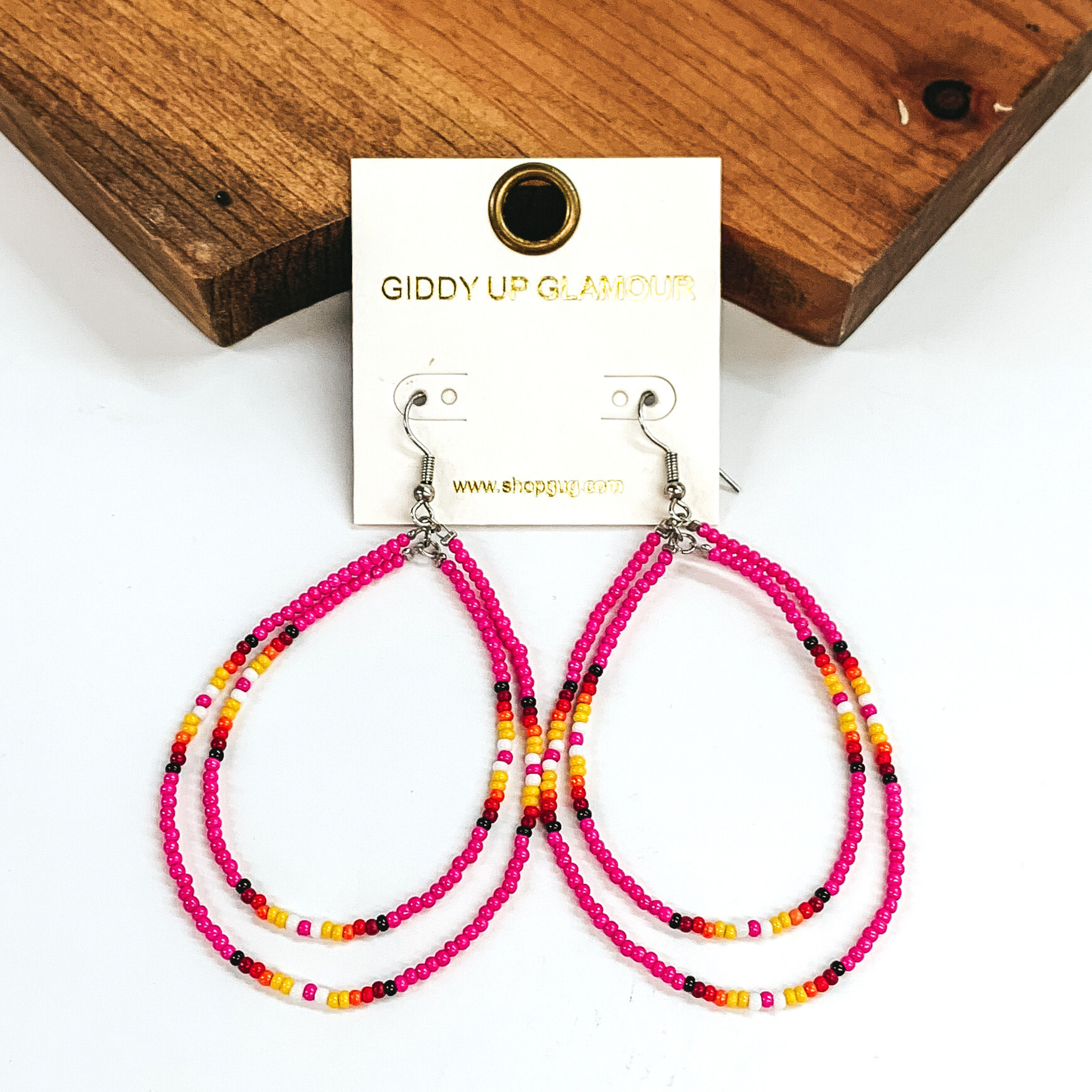 Double layered, hanging beaded teardrop earrings. The majority of the beads are pink with three segements including maroon, red, yellow, white, and black beads. hese earrings are pictured on a white background in front of a dark piece of wood. 