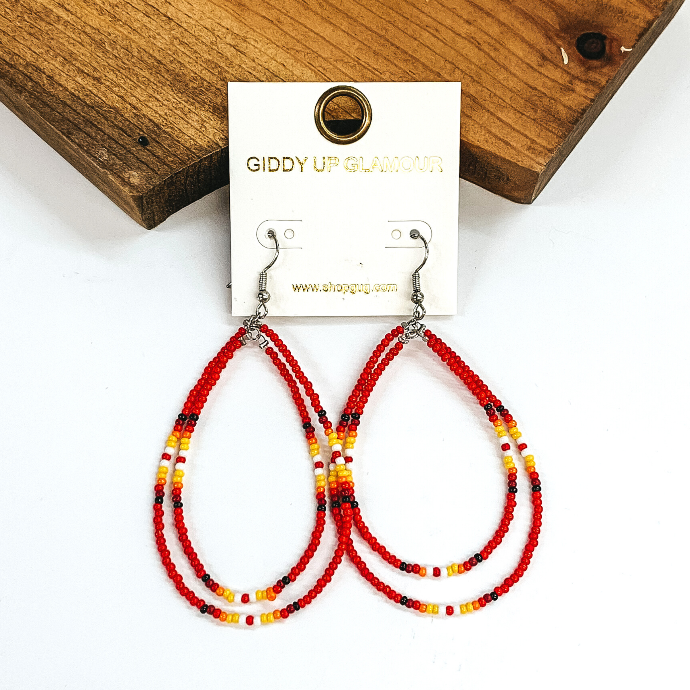 Double layered, hanging beaded teardrop earrings. The majority of the beads are red with three segements including maroon, red, yellow, white, and black beads. hese earrings are pictured on a white background in front of a dark piece of wood. 