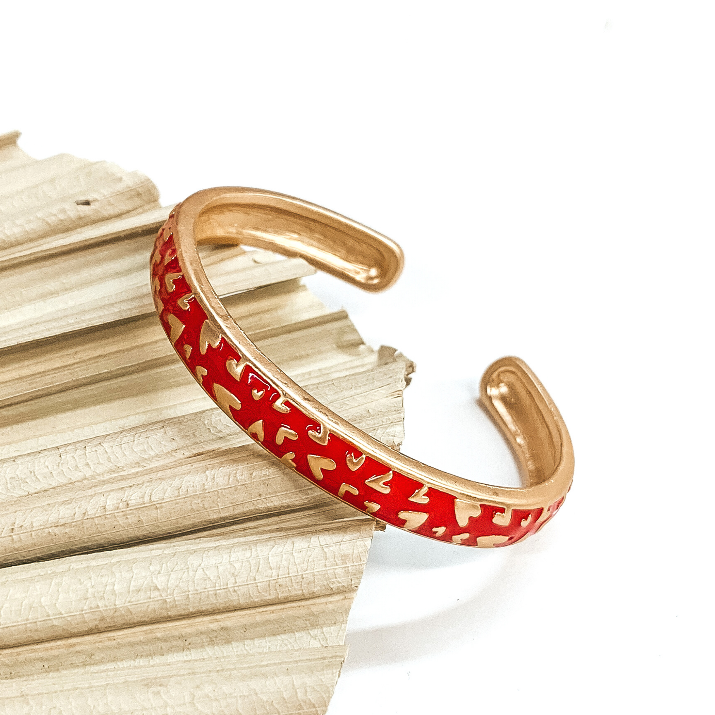 This is a gold outlined bracelet with a red coating and a gold heart pattern. This bracelet is pictured laying on a green palm leaf on a white background. 