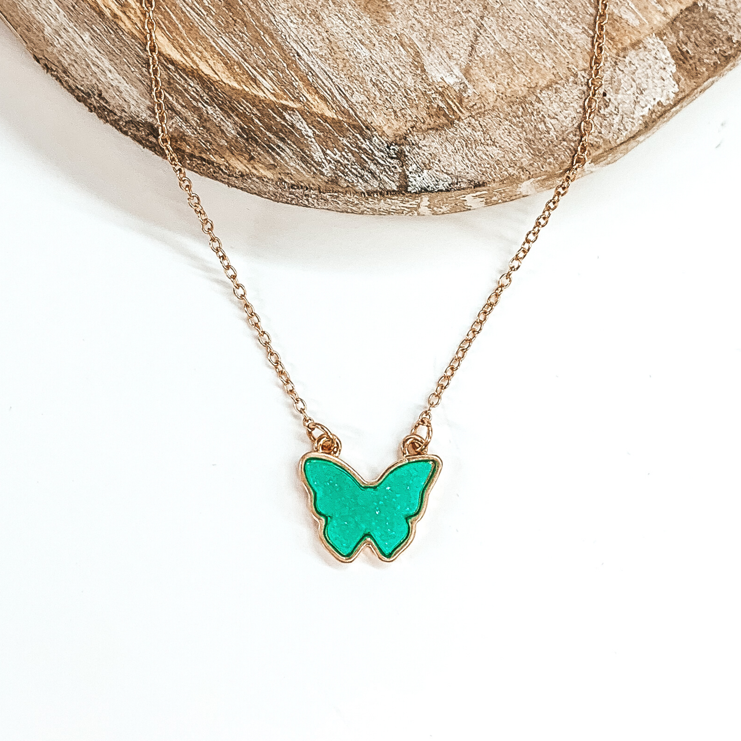 Tiny gold chained necklace includes a mint , druzy butterfly pendant that is outlined in gold. This necklace is pictured on a white background with a tan piece of wood at the top of the picture.