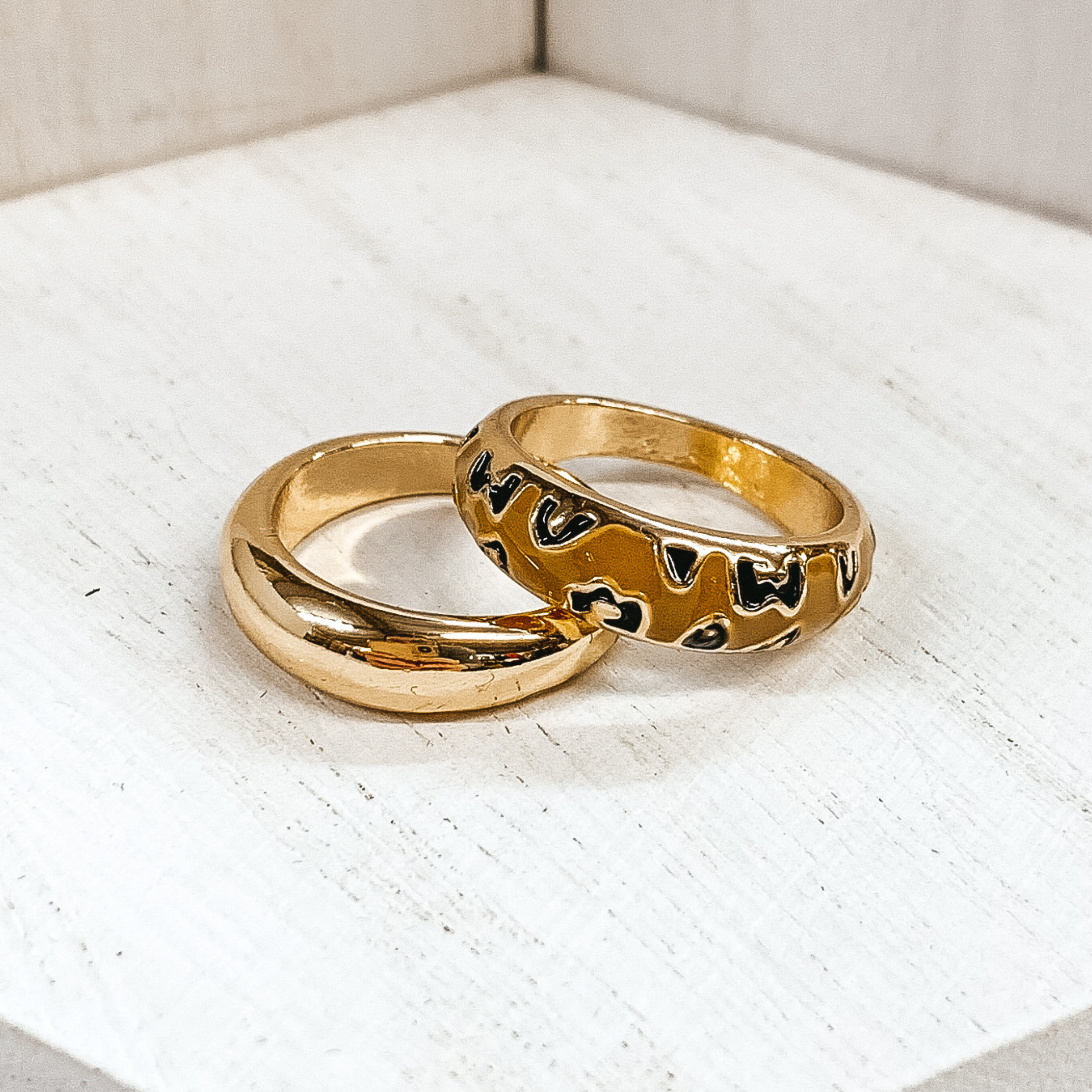 Set of two thick, gold rings. One ring is plain, while the secong ring has a tan colored part with leopard print. These rings are pictured on a white background. 