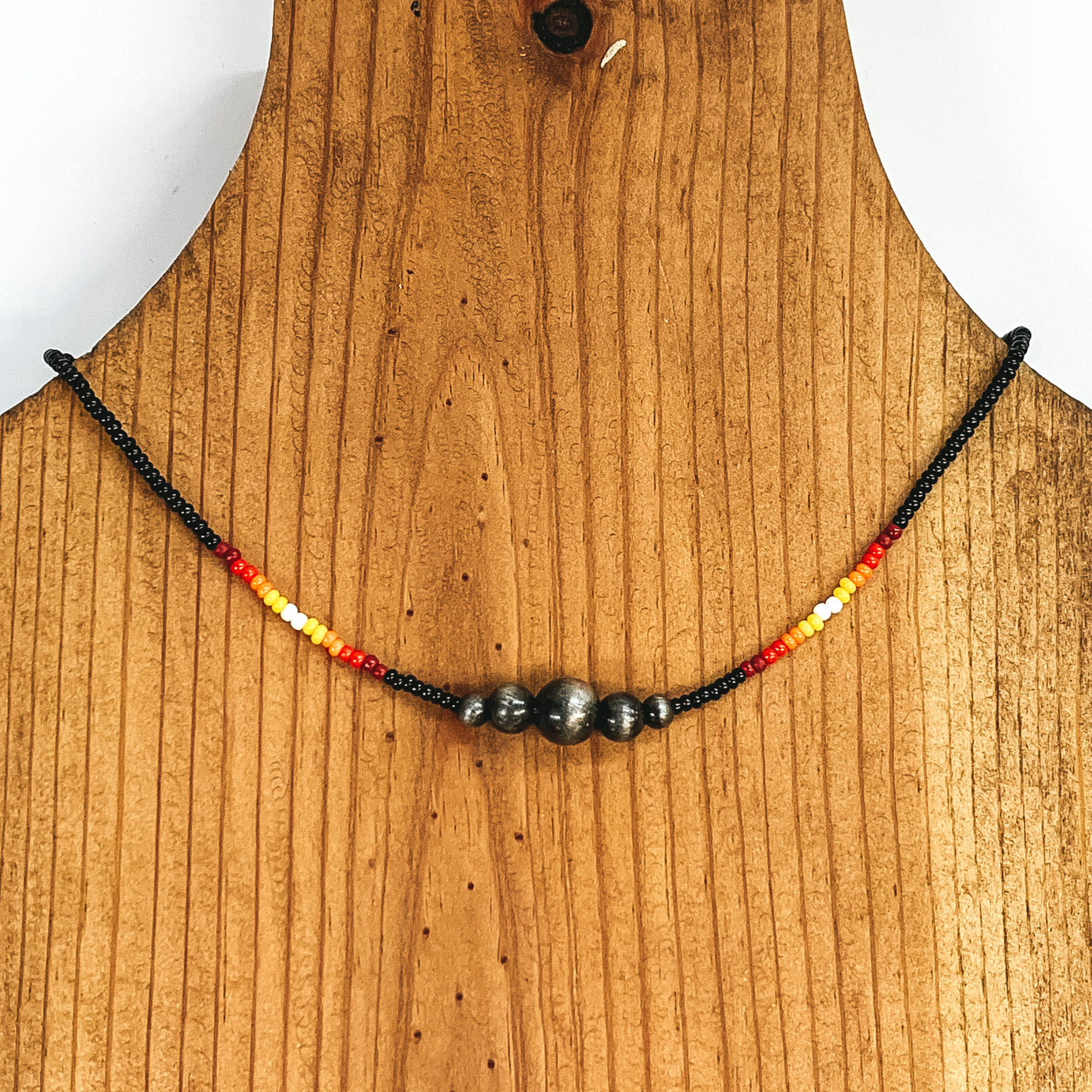 Black beaded necklace with center silver beads of varying sizes. On each side of the silver, center beads there is a segment the has white center beads with yellow, orange, red, and maroon beads folowing on each side. This necklace is pictured on a wooden necklace holder on a white background. 