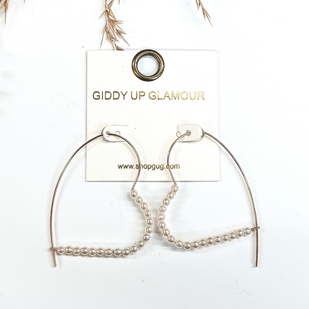 Kidney thin wire earrings shaped in a heart  in gold with small pearl beads in the bottom half. Taken ina white background with a brown plant in the  back as decor.