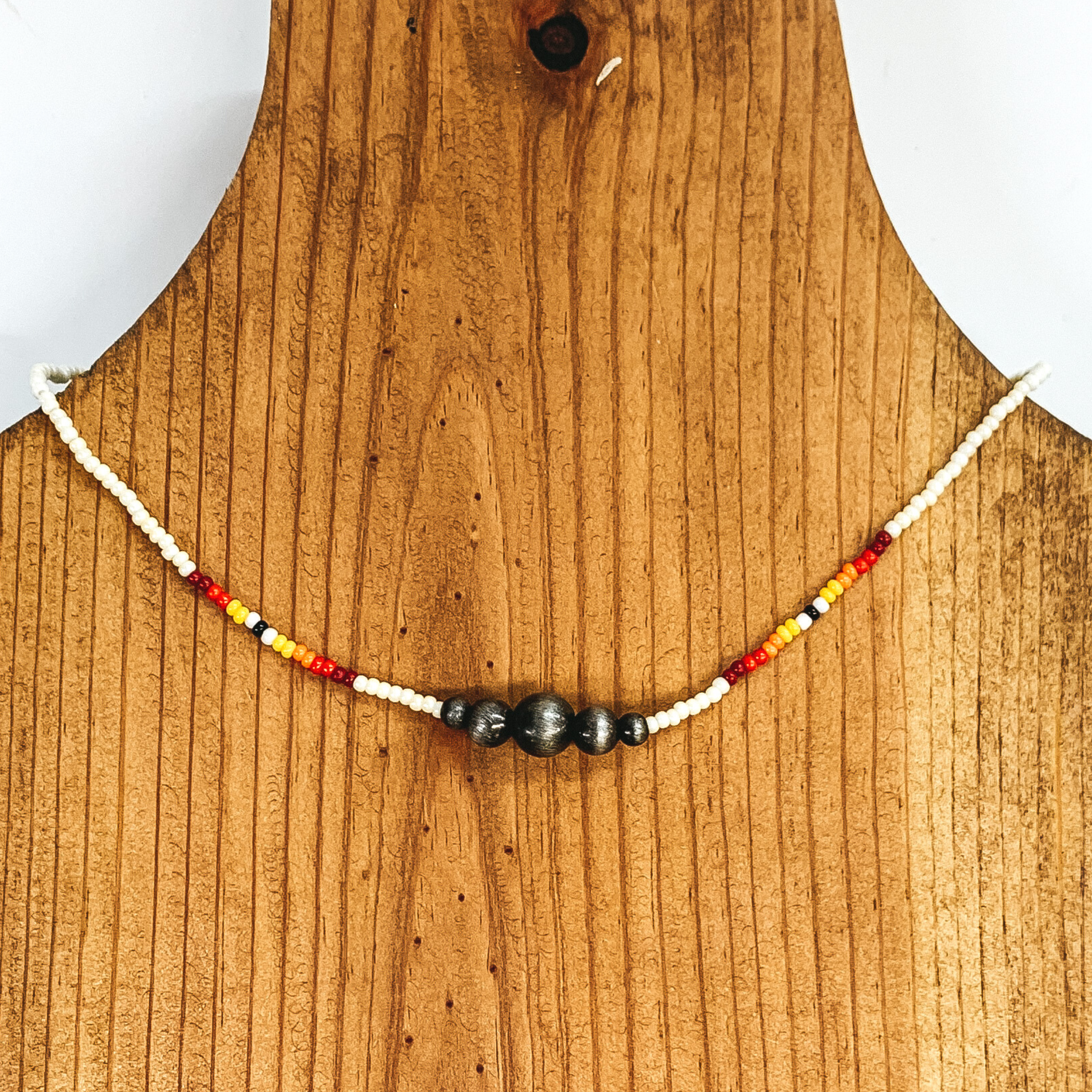 White beaded necklace with center silver beads of varying sizes. On each side of the silver, center beads there is a segment the has black center bead with white, yellow, orange, red, and maroon beads folowing on each side. This necklace is pictured on a wooden necklace holder on a white background. 