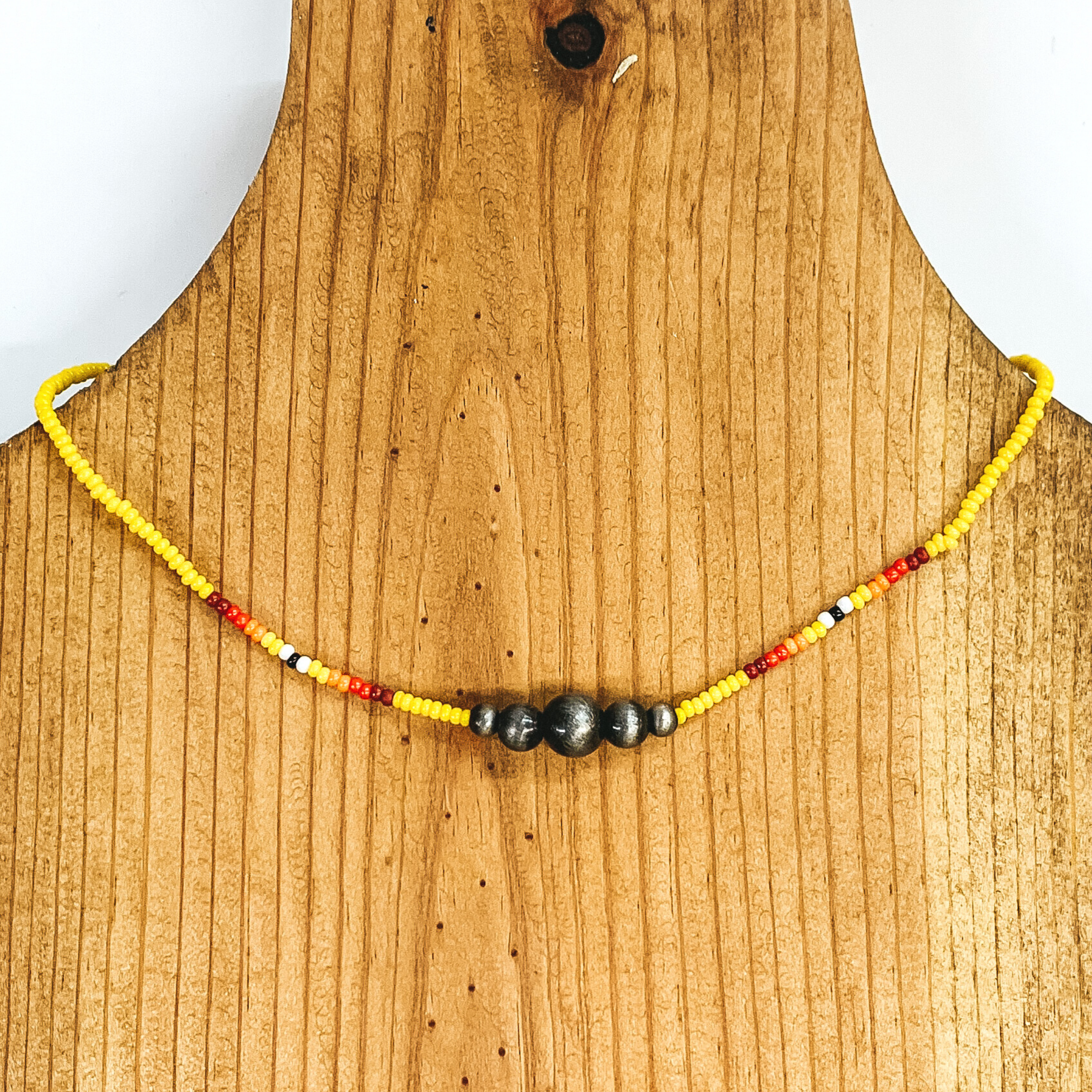Yellow beaded necklace with center silver beads of varying sizes. On each side of the silver, center beads there is a segment the has black center bead with white, yellow, orange, red, and maroon beads folowing on each side. This necklace is pictured on a wooden necklace holder on a white background. 