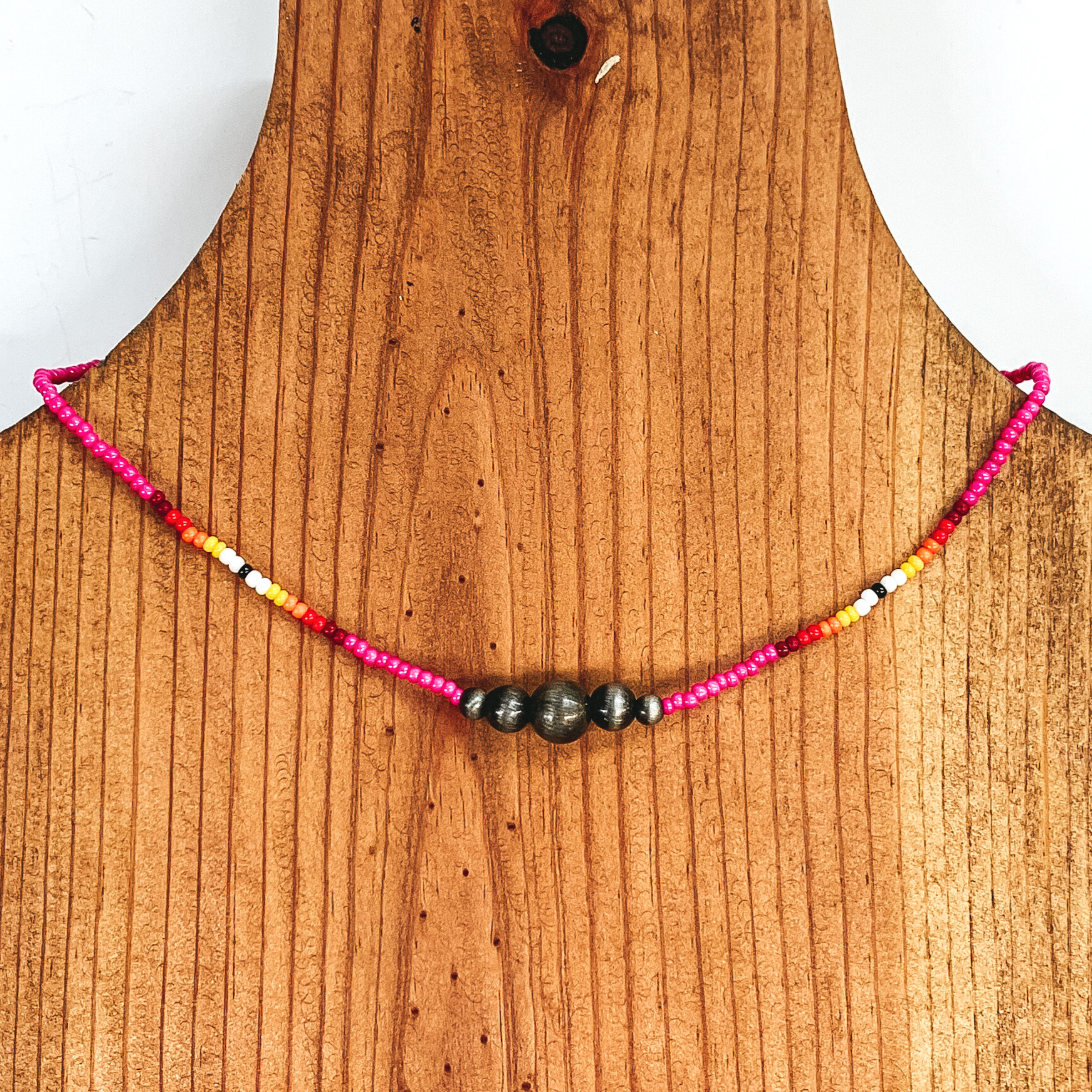 Pink beaded necklace with center silver beads of varying sizes. On each side of the silver, center beads there is a segment the has black center bead with white, yellow, orange, red, and maroon beads folowing on each side. This necklace is pictured on a wooden necklace holder on a white background. 