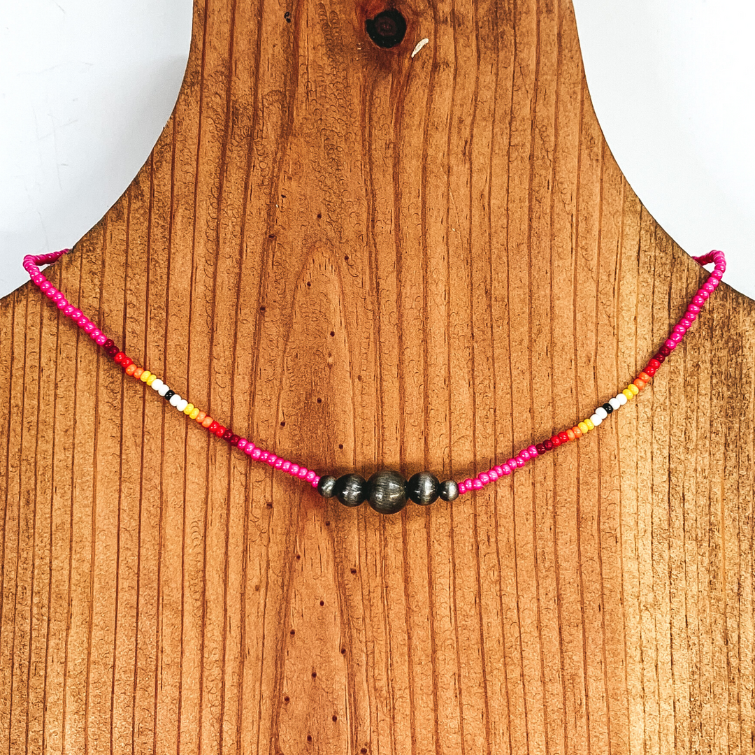 Pink beaded necklace with center silver beads of varying sizes. On each side of the silver, center beads there is a segment the has black center bead with white, yellow, orange, red, and maroon beads folowing on each side. This necklace is pictured on a wooden necklace holder on a white background. 