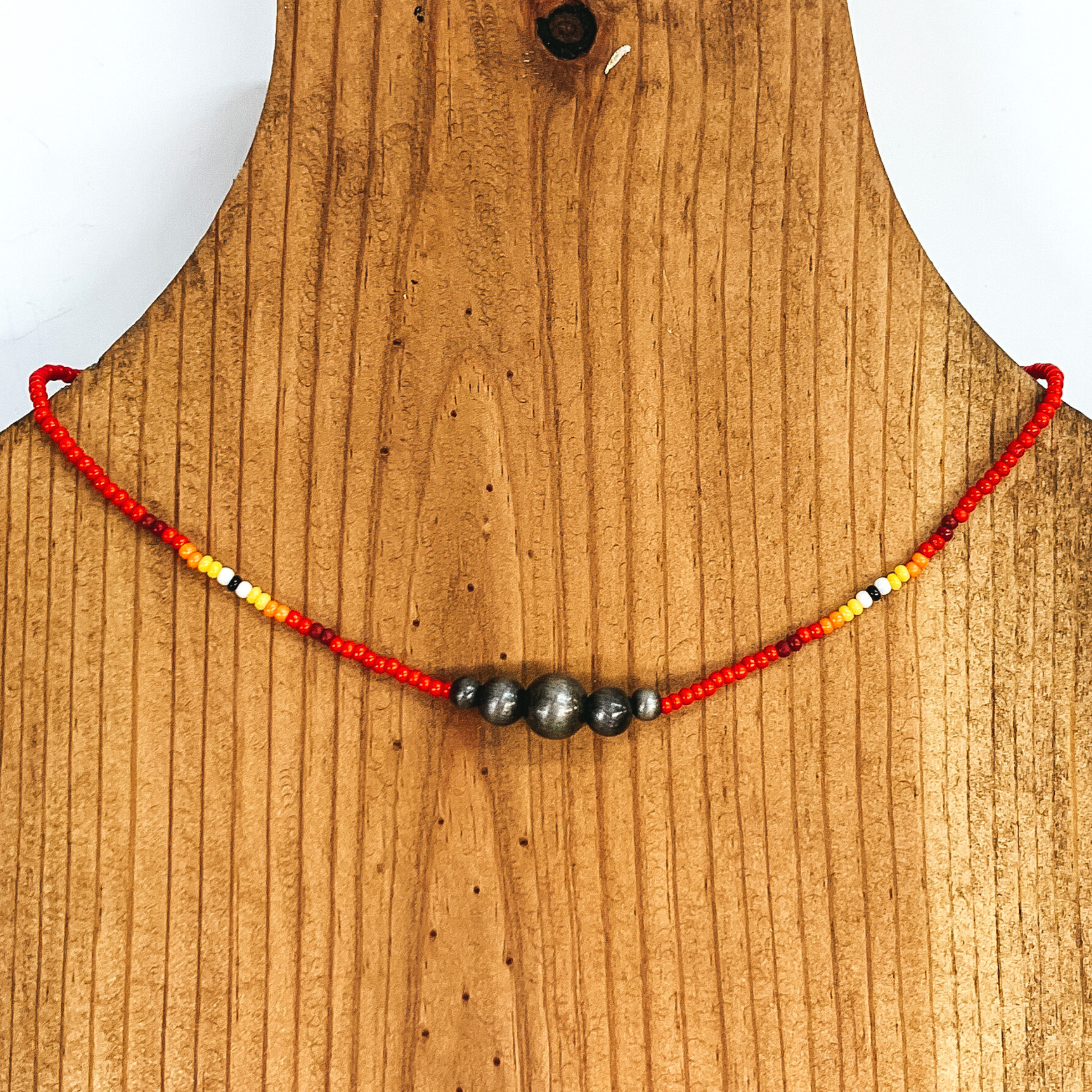 Red beaded necklace with center silver beads of varying sizes. On each side of the silver, center beads there is a segment the has black center bead with white, yellow, orange, red, and maroon beads folowing on each side. This necklace is pictured on a wooden necklace holder on a white background. 