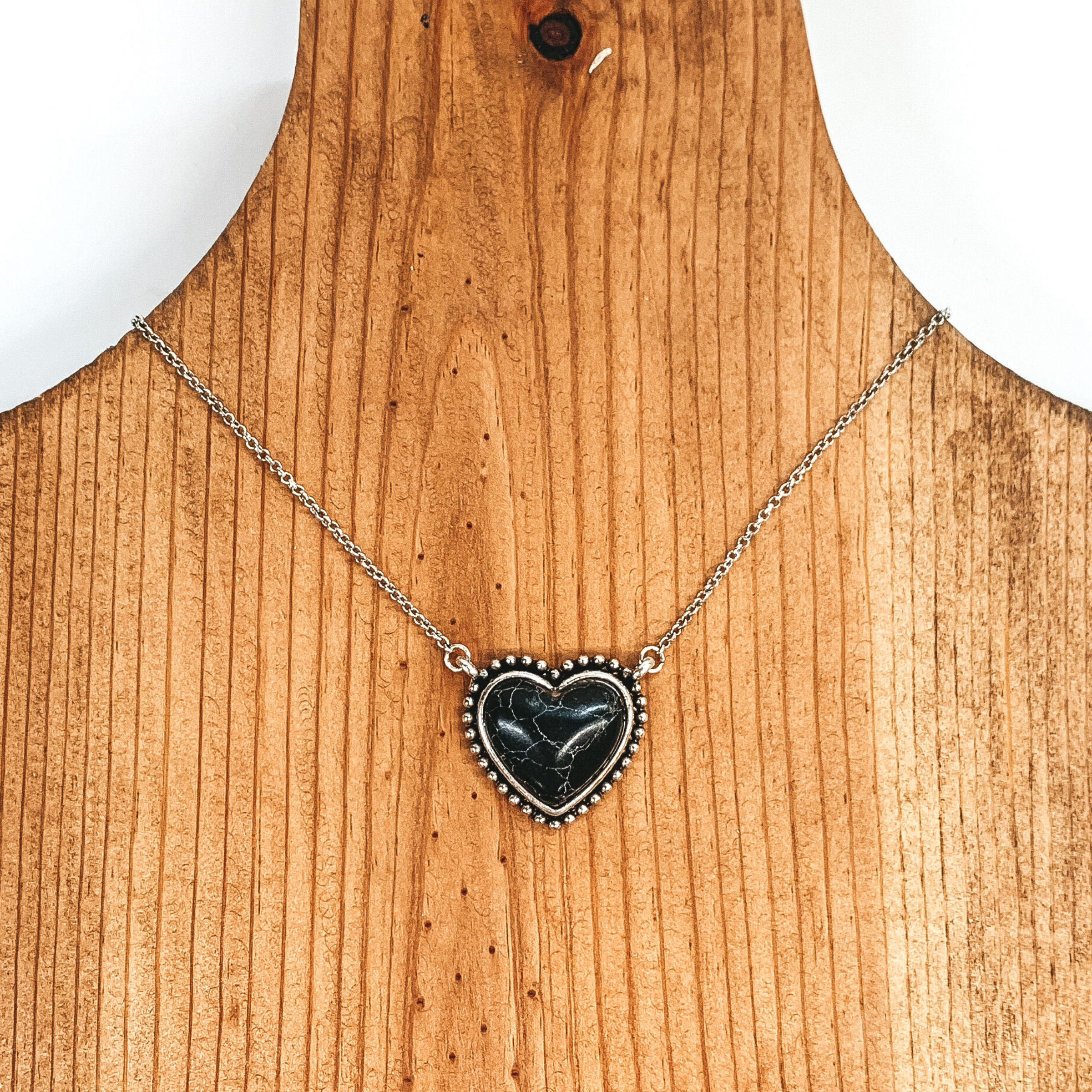 Silver chained necklace with a small black, heart shaped stone pendant. This necklace is pictured laying on a brown necklace holder on a white and black background.