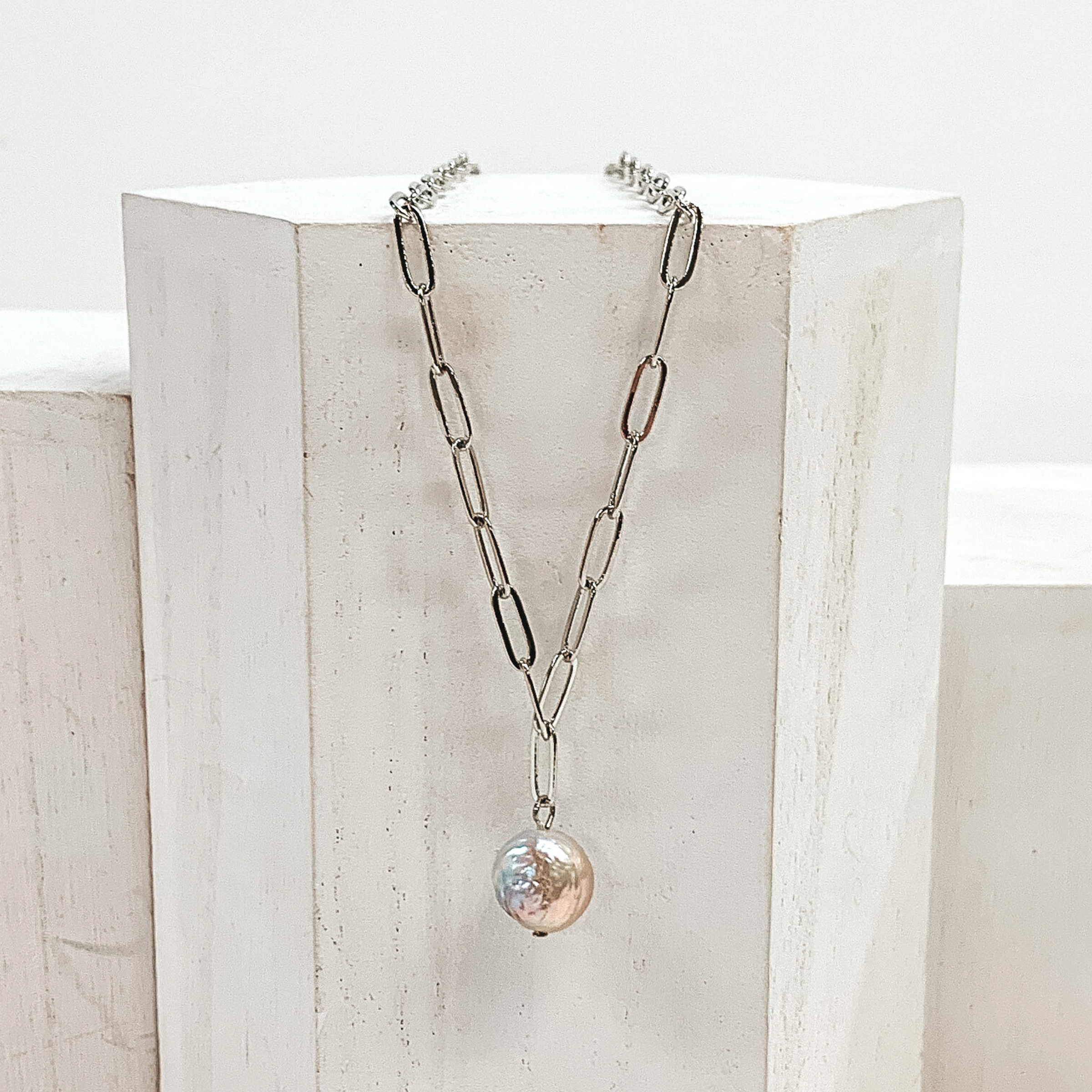 This is a silver paperclip chained necklace with a irregularly shaped pearl drop pendant. This necklace is pictured laying on a white block on a white background.