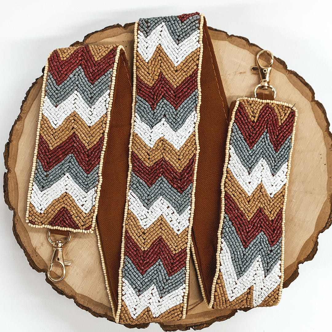 This purse strap has a brown, grey, and white chevron striped pattern with khaki colored backing. This purse strap has gold clasps. This purse strap is pictured on a piece of wood on a white background.