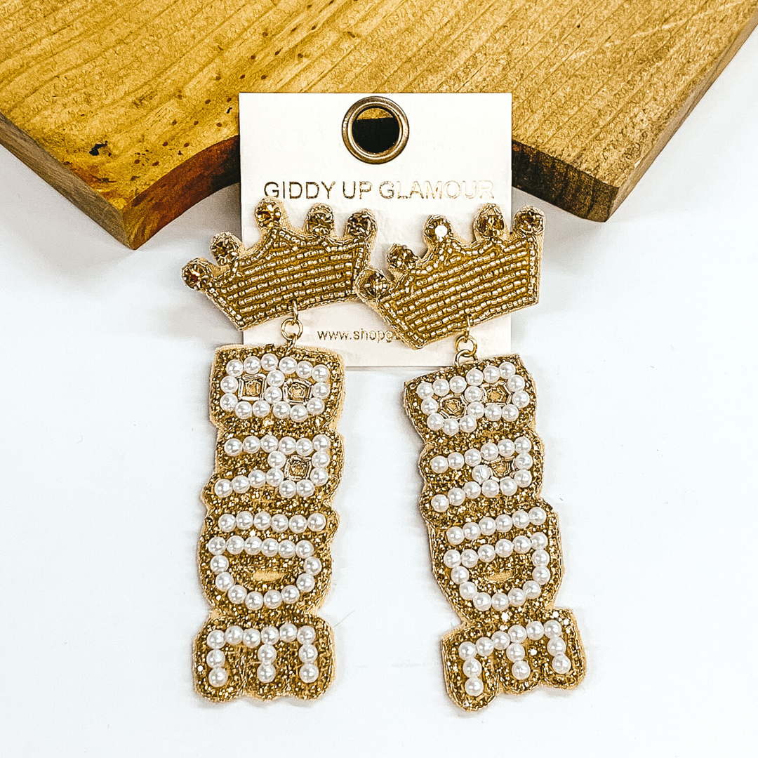 Gold beaded crown stud earrings with gold crystals. These crown studs have a hanging beaded "BRIDE" in gold crystals and white beads. These earrings are pictured on a white earrings holder laying against a dark brown block on a white background.