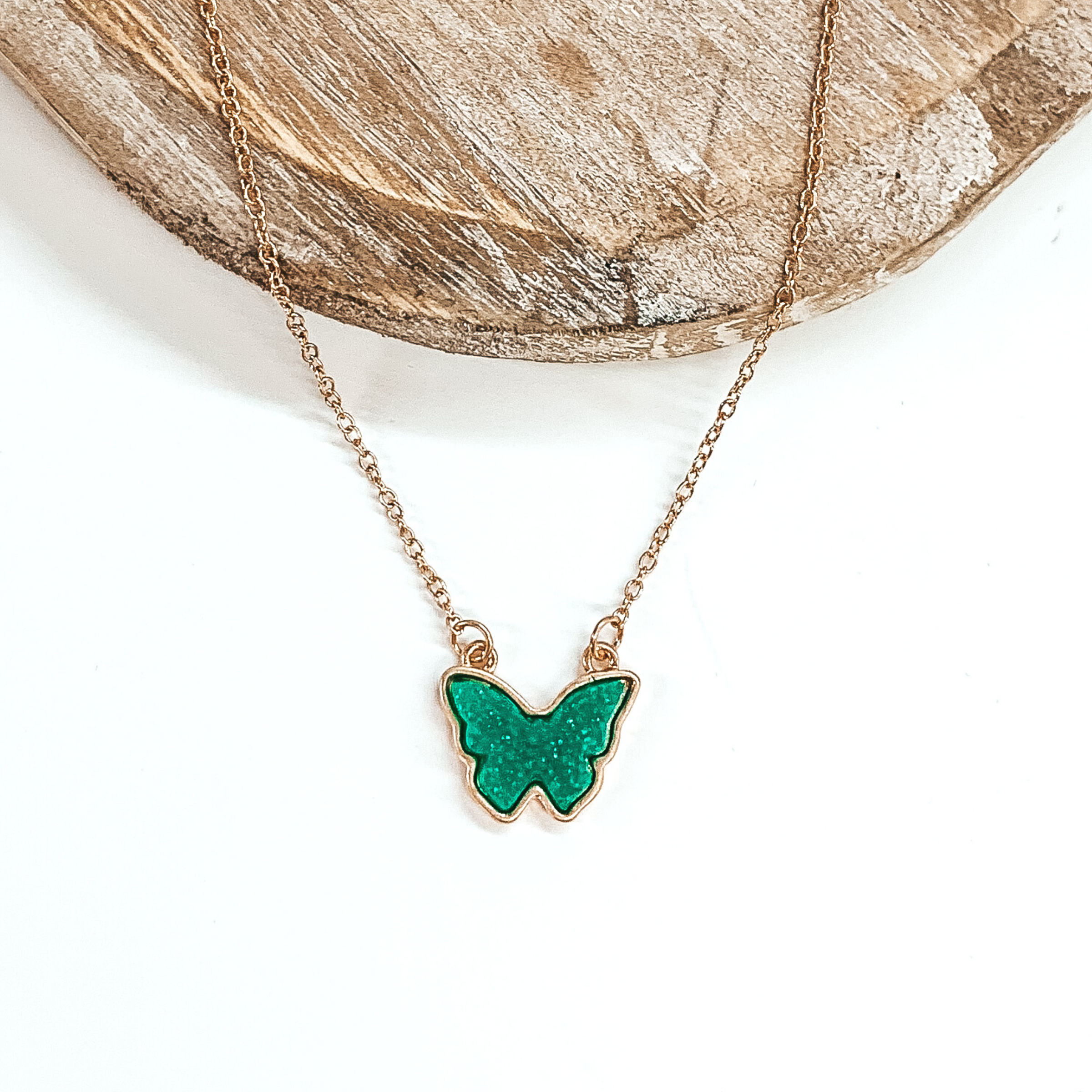 Tiny gold chained necklace includes a turquoise, druzy butterfly pendant that is outlined in gold. This necklace is pictured on a white background with a tan piece of wood at the top of the picture.