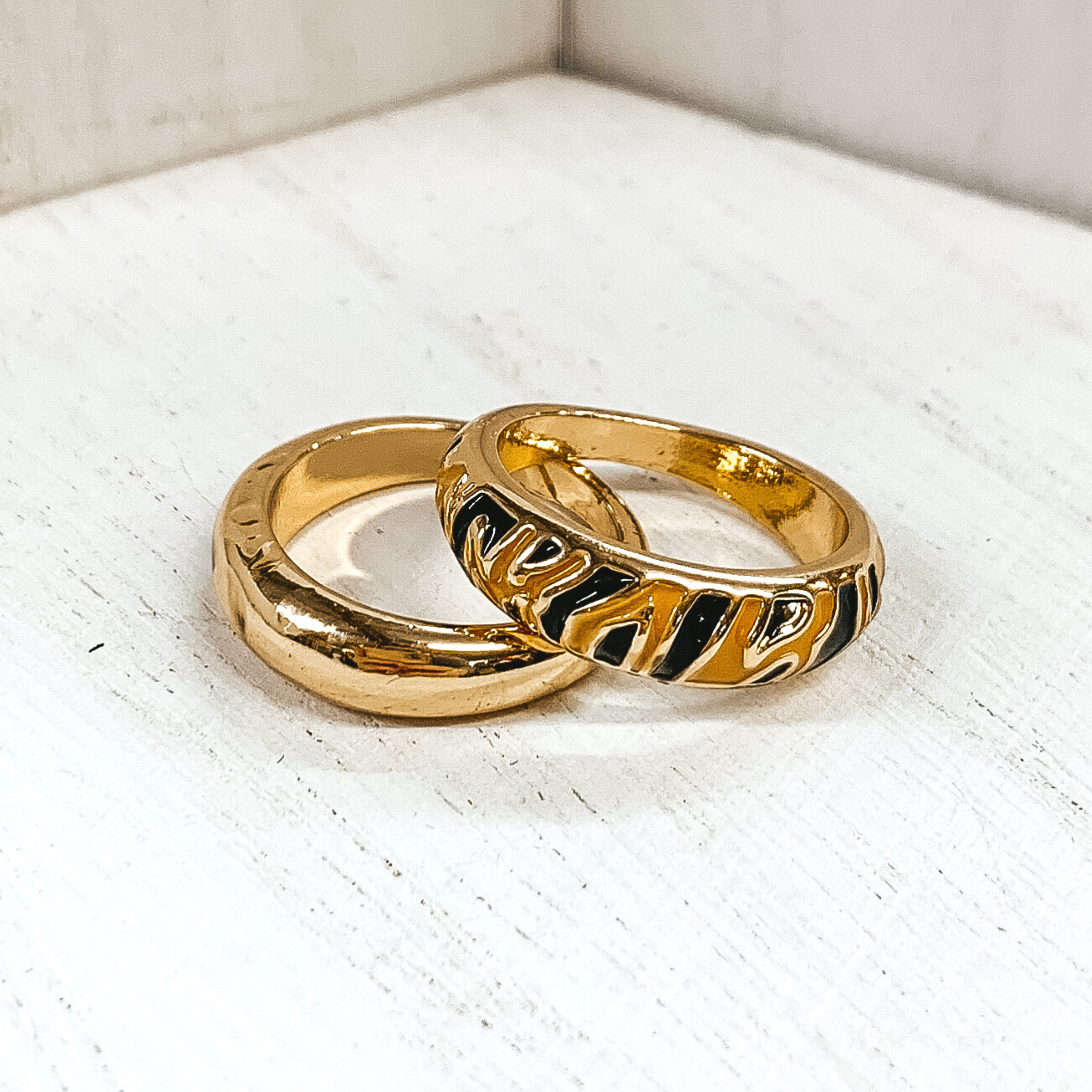 Set of two thick, gold rings. One ring is plain, while the secong ring has a tan colored part with tiger stripes. These rings are pictured on a white background. 